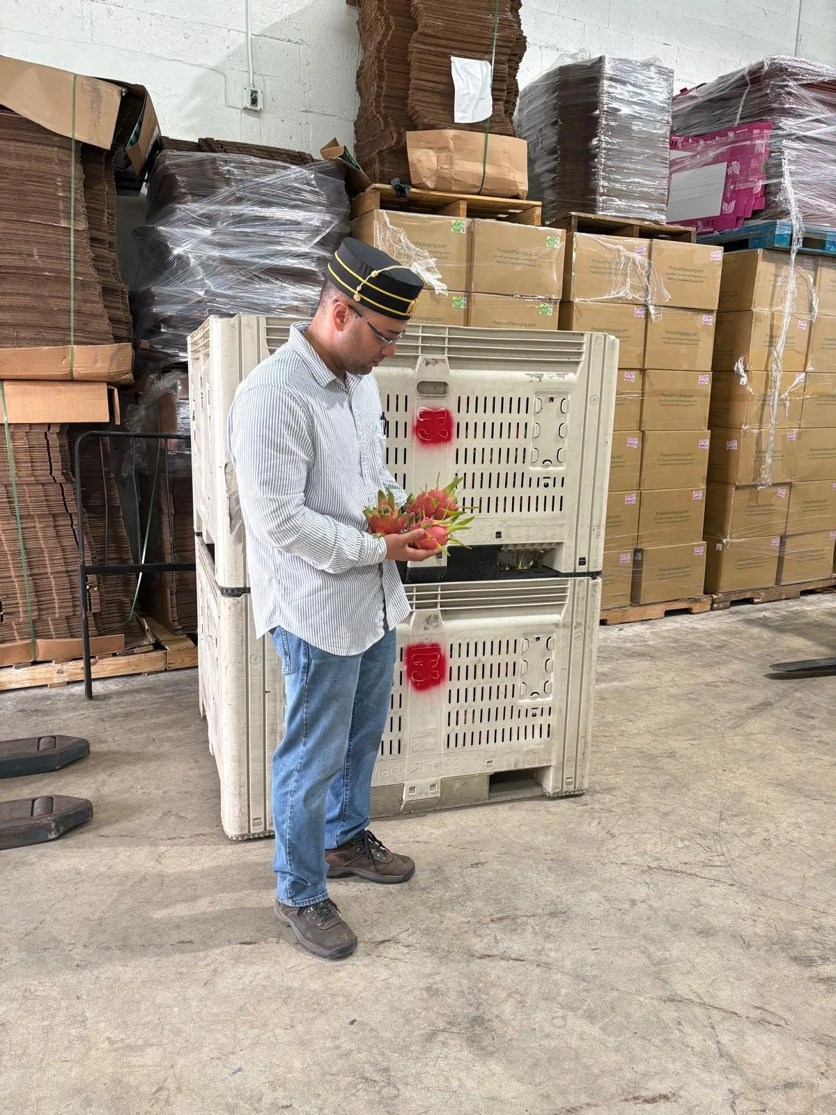 A man wearing a striped hat and glasses stands in a warehouse holding a bunch of pink flowers. Behind him are stacked cardboard boxes, wrapped pallets, and plastic containers, with a concrete wall and floor.