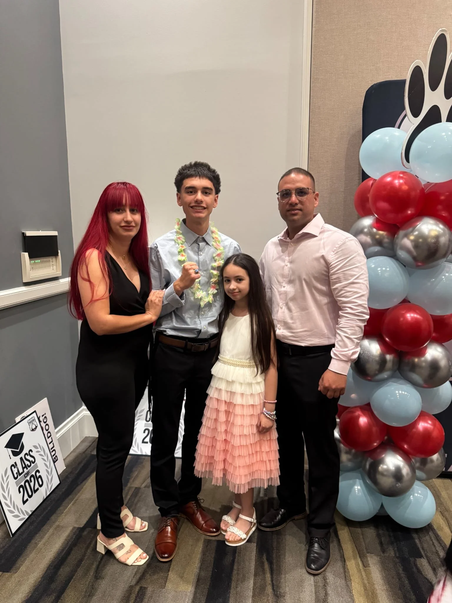 A group of five people standing together at a graduation celebration, with balloons and signs visible in the background.