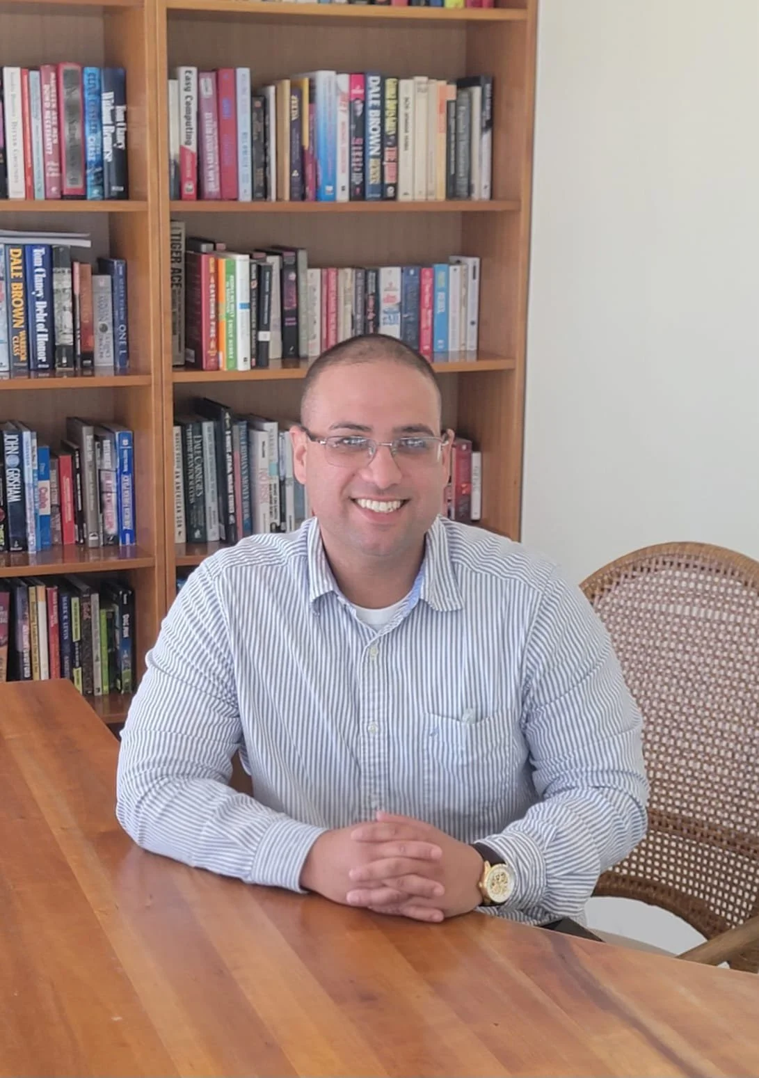 A man with glasses and a striped shirt sitting at a wooden table in front of a bookshelf filled with books, smiling at the camera.