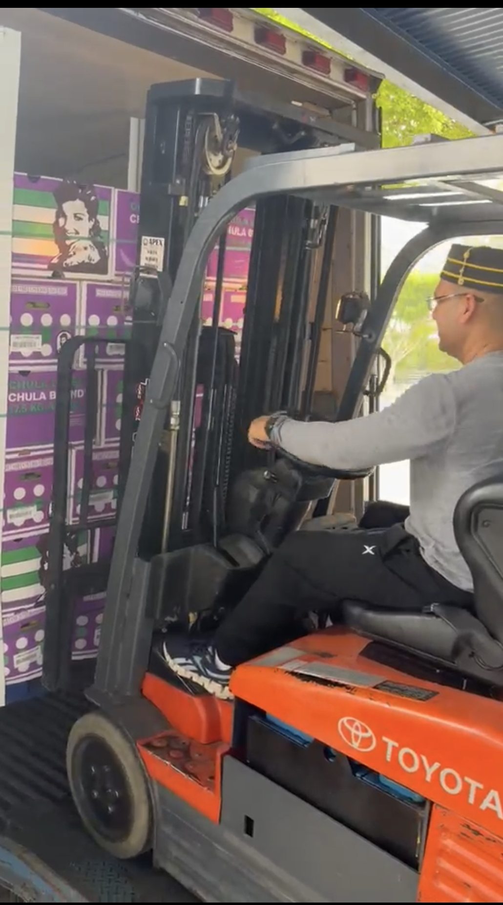 A man wearing a gray shirt and a sailor hat operates an orange Toyota forklift to move purple boxes with a poster of a woman’s face on the side.