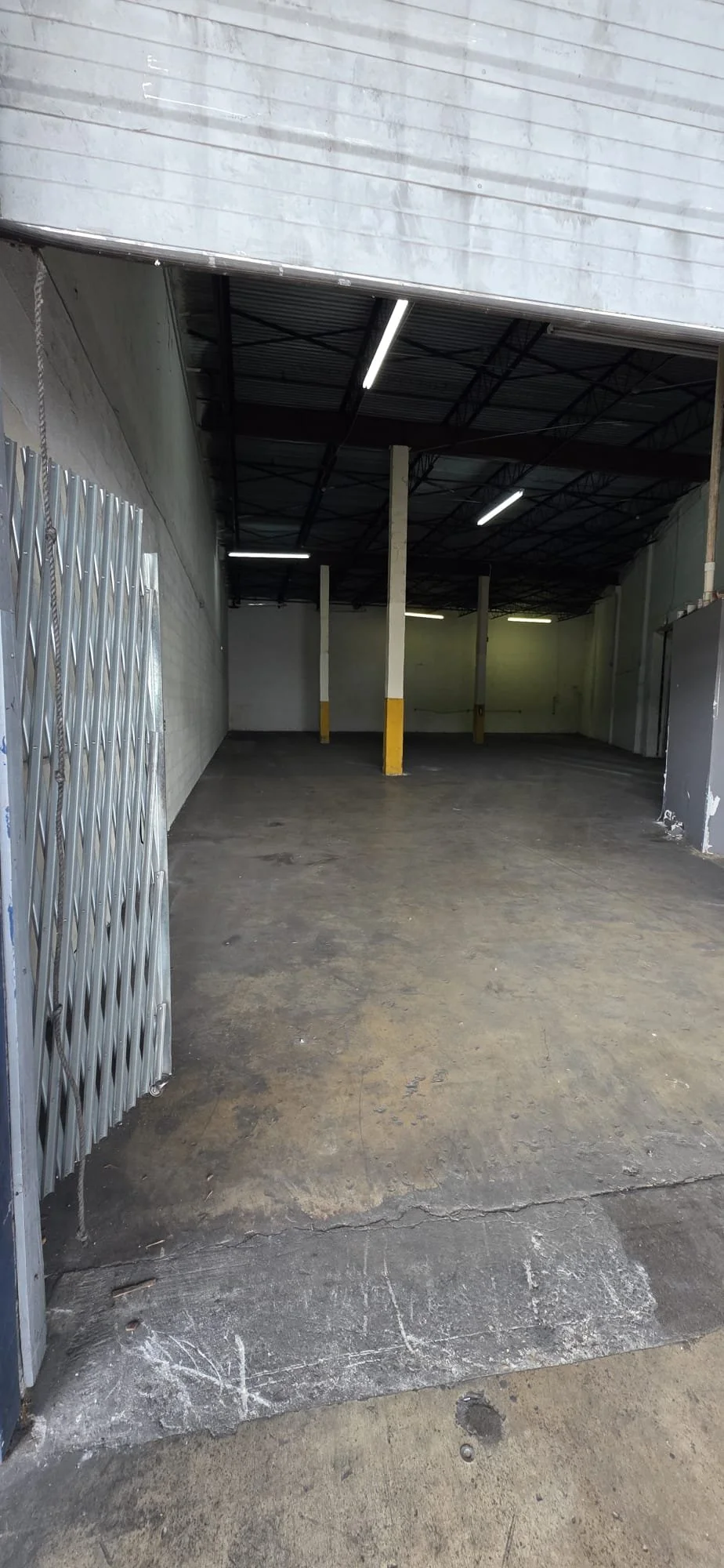 Empty warehouse or garage with concrete floor, white walls, drop ceiling, and fluorescent lights. There are metal accordion doors partially open on the left side.