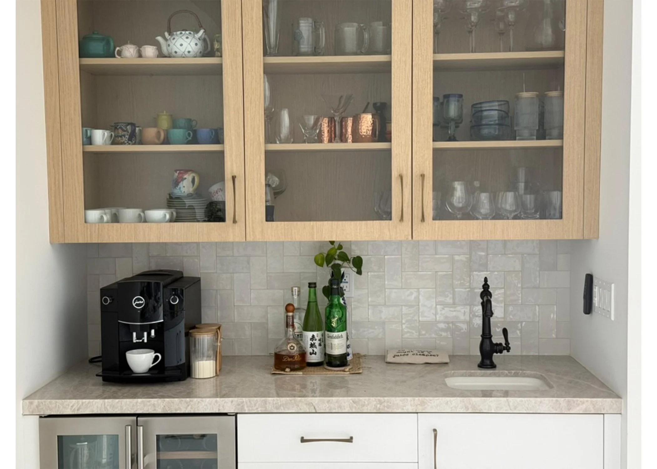A kitchen countertop with a black coffee maker, a white mug, a small container of milk, and various bottles on a tray. Behind the bottles are a paper towel roll and a cutting board. A black faucet with a sink is on the right, and lower cabinets are also visible.