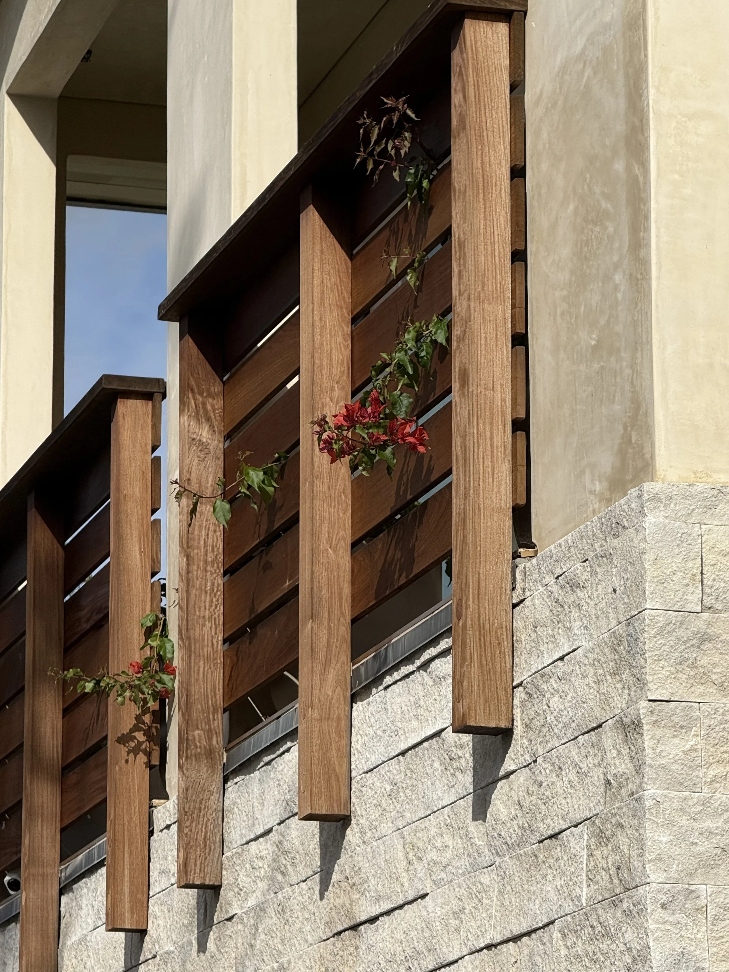 A wooden balcony railing attached to a beige stucco building with stone foundation, with green and red plants climbing on the railing.