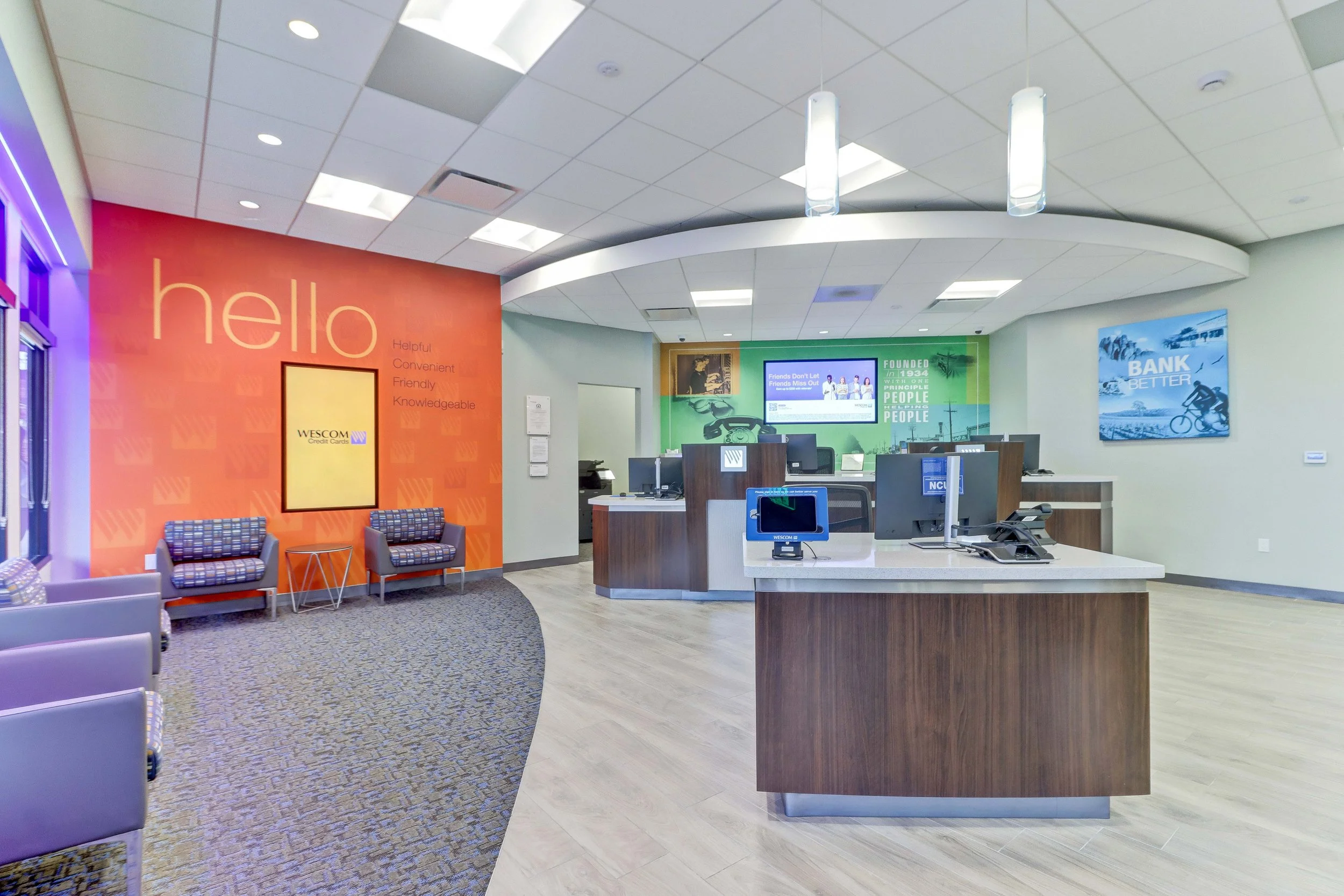 A modern bank interior with a waiting area on the left, featuring three chairs and a small table, and a service counter on the right with computers and monitors. The background has colorful wall decorations, including a large orange wall with the wor
