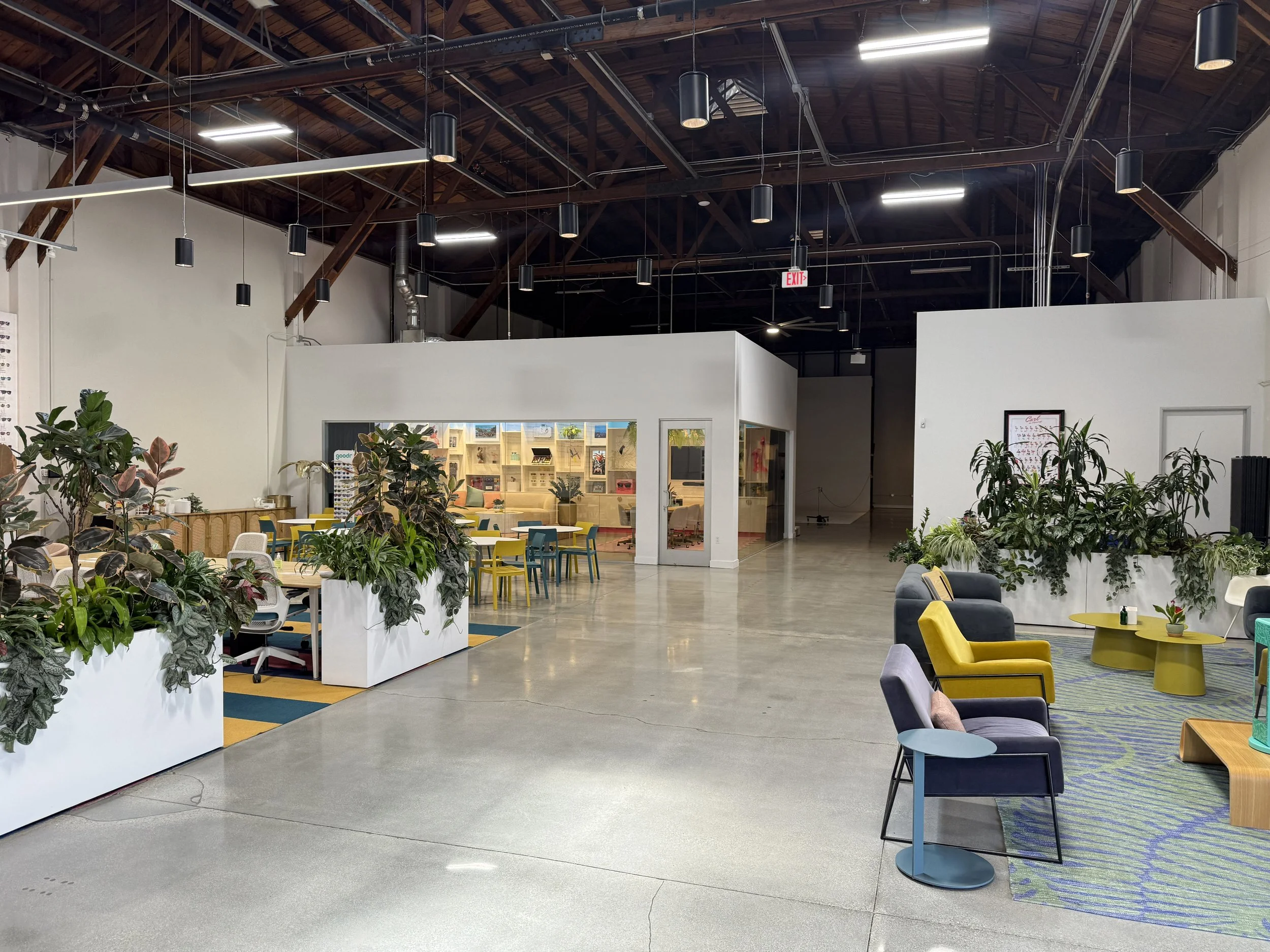 Open indoor space with high wooden ceiling, hanging pendant lights, and various colorful chairs and tables, some with plants and decorations.