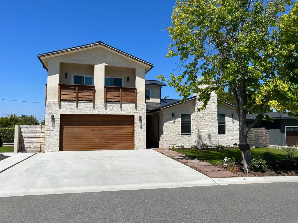Modern suburban house with a two-car garage, wooden balcony, brick pathways, a large tree, and a well-maintained lawn under a clear blue sky.