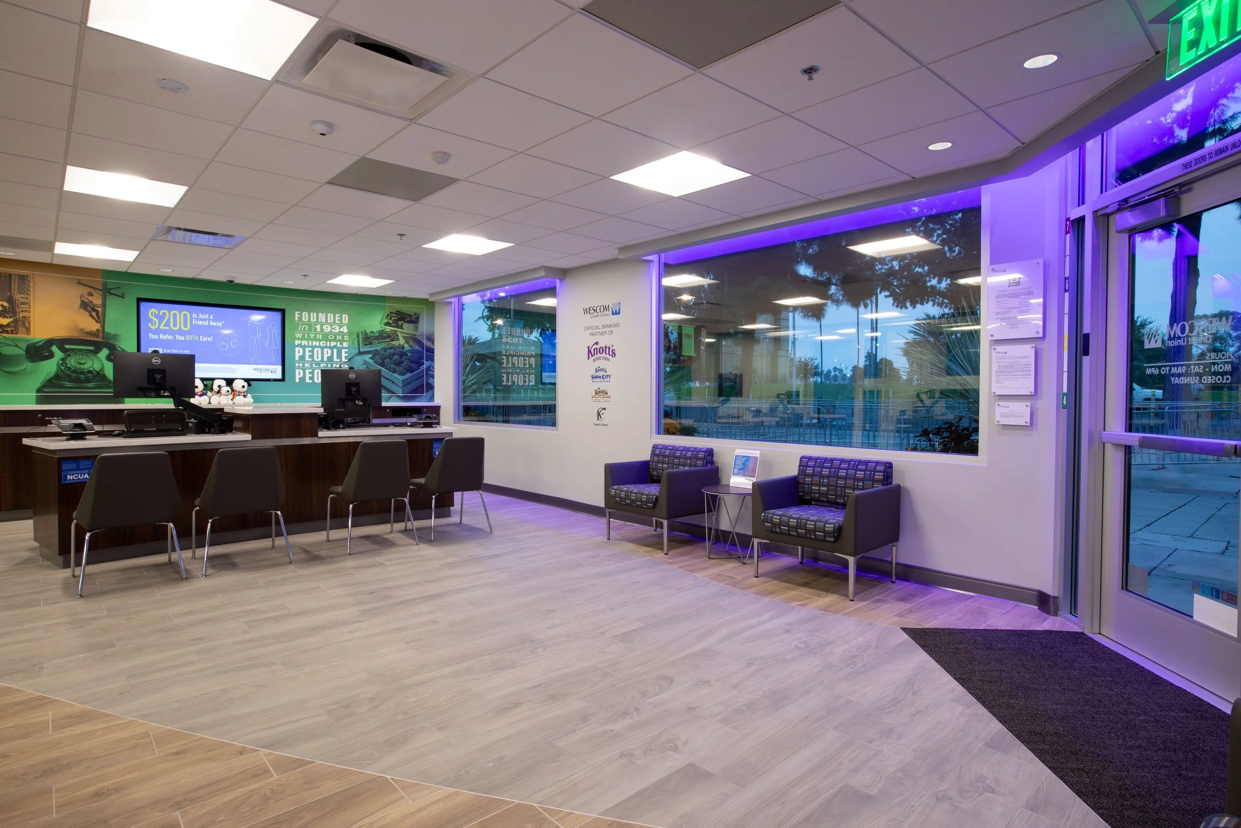 Interior of a bank or reception area with check-in counters, waiting chairs, and large windows outside, illuminated by ceiling lights and purple LED lighting.