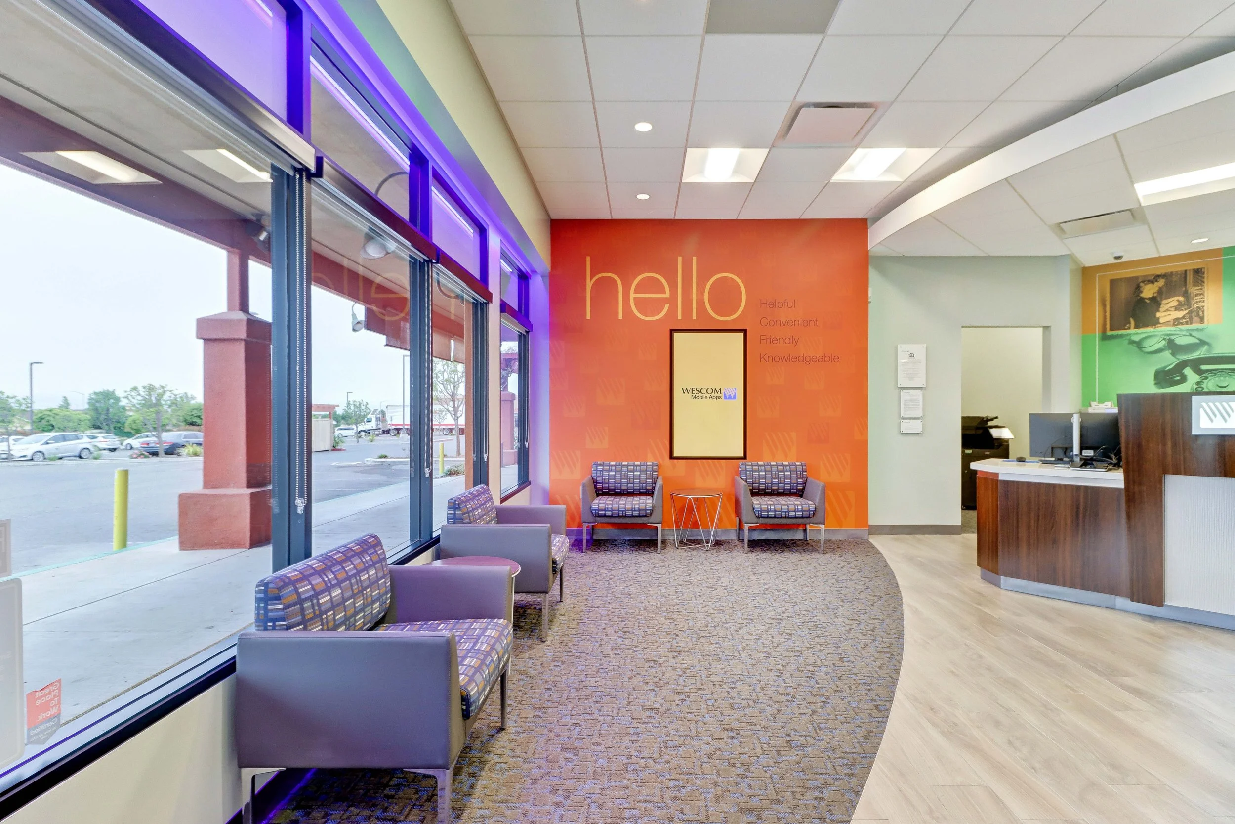 Inside of a modern building with a waiting area near large glass windows. The wall features the word 'hello' in large letters, along with additional friendly descriptive words. There are chairs with striped upholstery and a small table. The ceiling has a grid pattern with recessed lighting, and a reception desk is visible on the right. Outside, a parking lot can be seen through the windows.