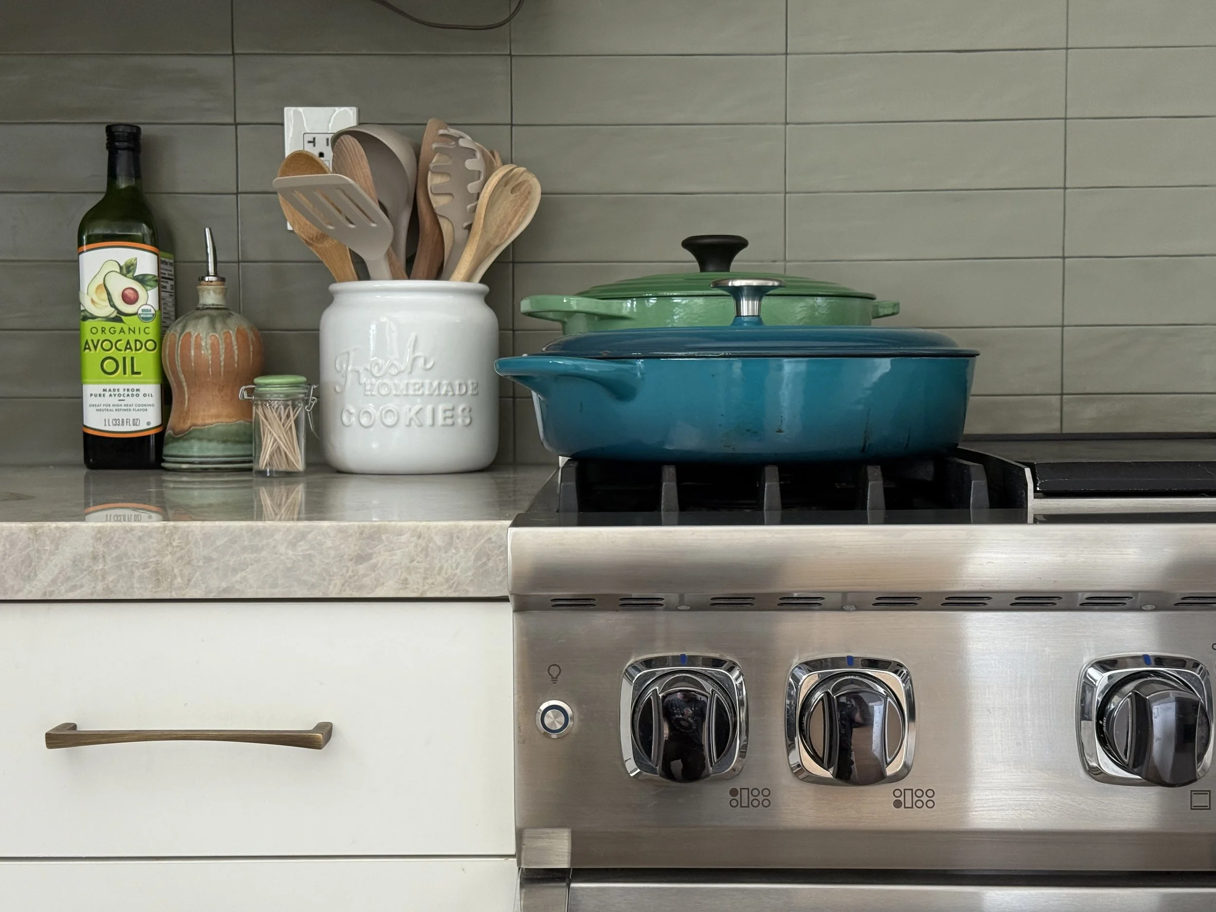 Kitchen countertop with cooking pots, utensils in a container, avocado oil, jars, and a stovetop.