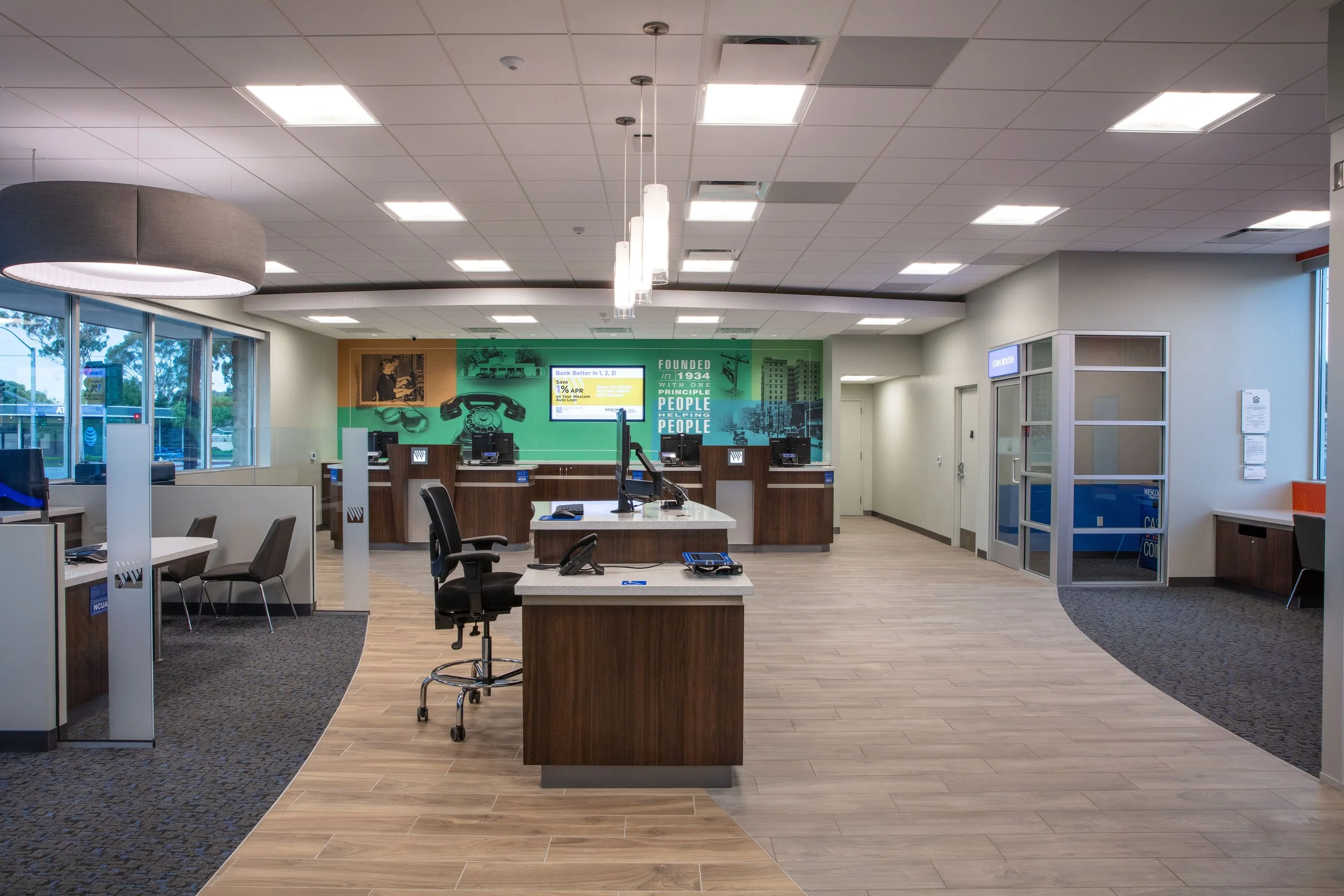 Empty bank lobby with countertops, chairs, and a green wall with a promotional sign, large windows on the left, and a glass-enclosed office area on the right.