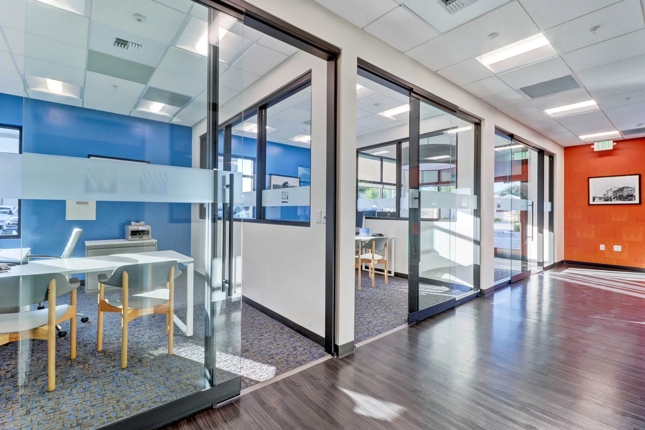 Interior view of an office with glass walls, multiple windows, and a mix of carpeted and wood flooring. There are office chairs, desks, and framed artwork visible on the walls.