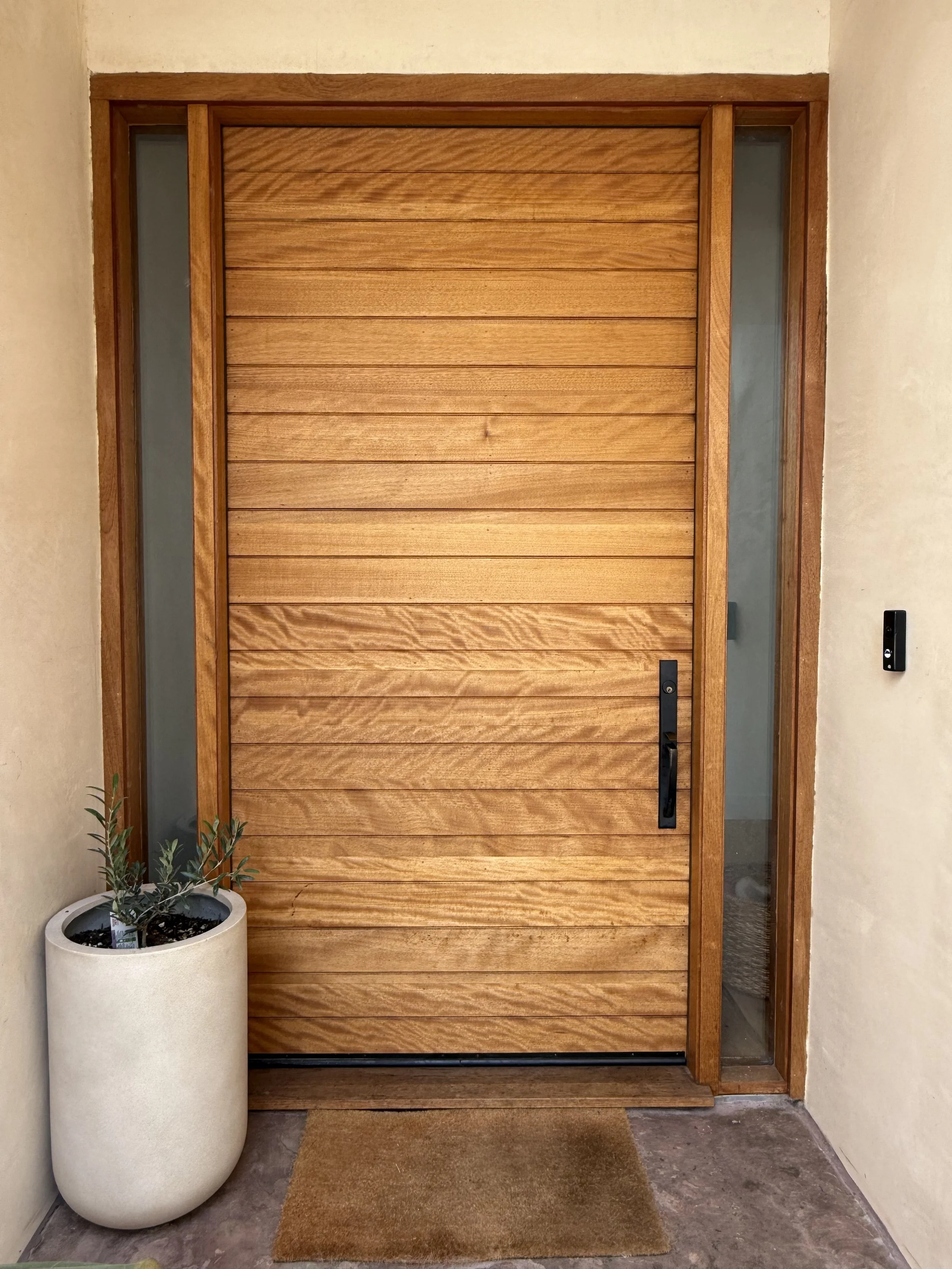 Front door made of horizontal wooden planks, with glass sidelight on each side and a black handle. A potted plant in a tall white planter is on the left, and a doormat is in front of the door on a concrete floor.