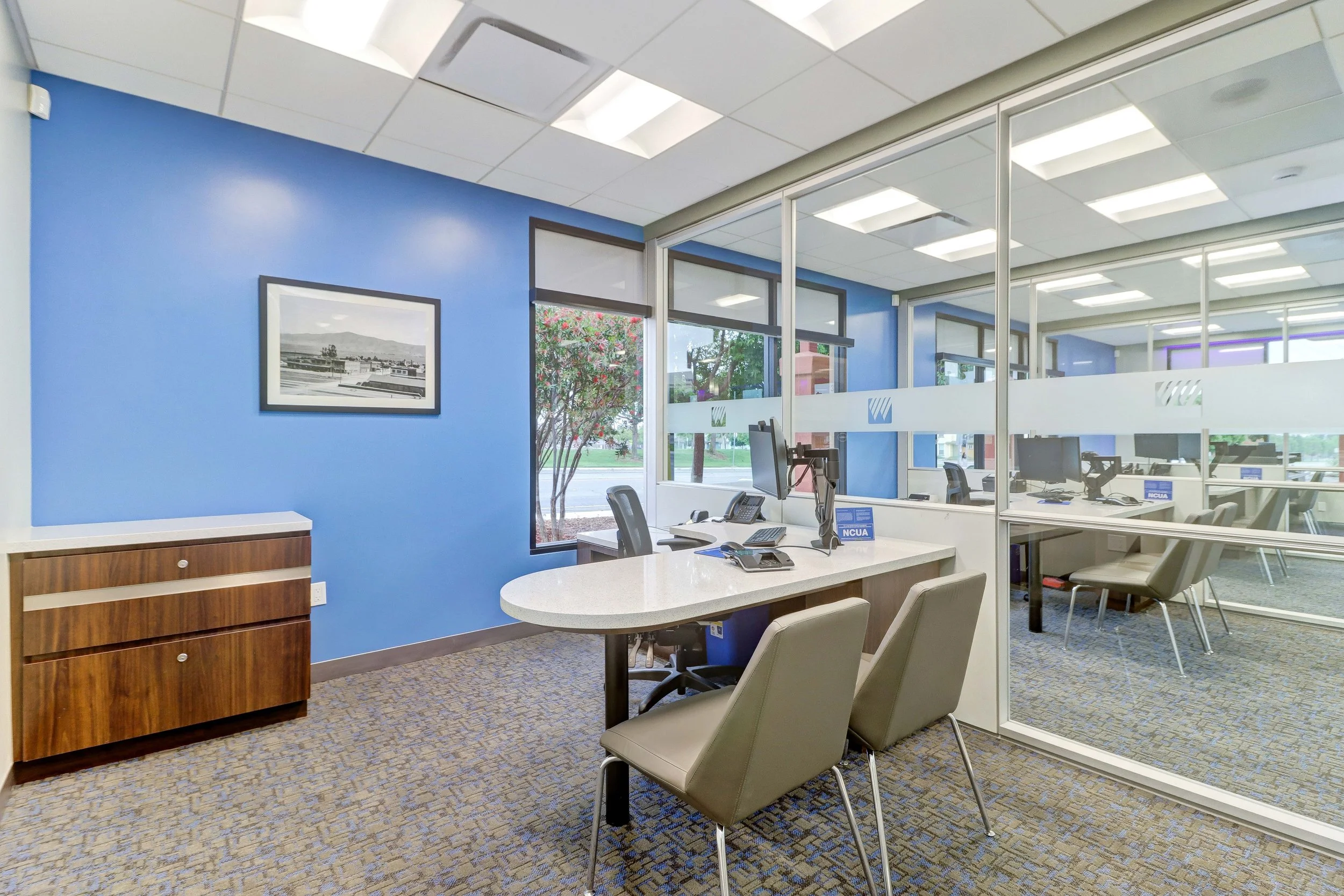 Office reception area with blue accent wall, framed picture, wooden cabinet, and white round table with chairs. Office desks visible through glass partition, with computer monitors and signs reading NCUA.