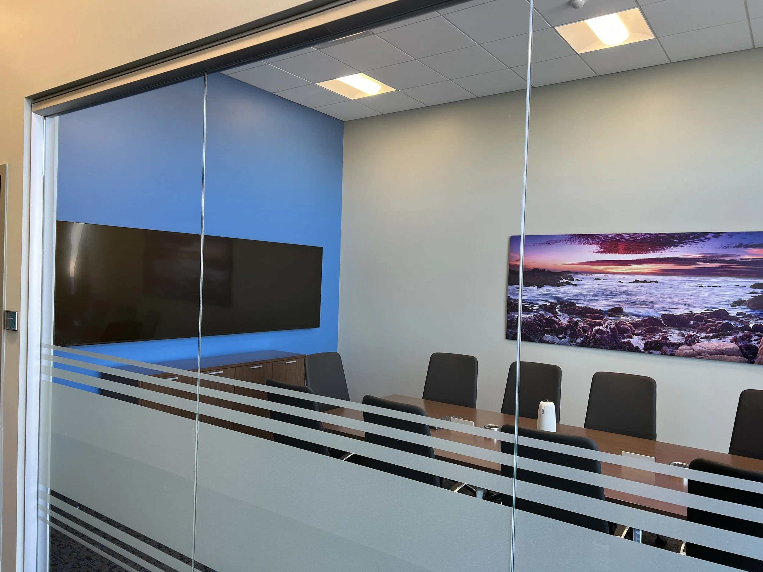 A conference room with glass walls and horizontal stripe decals. Inside, there is a large flat-screen TV on a blue wall, a long table with black chairs, and a landscape photo of a rocky beach at sunset hanging on a white wall. The ceiling has square 