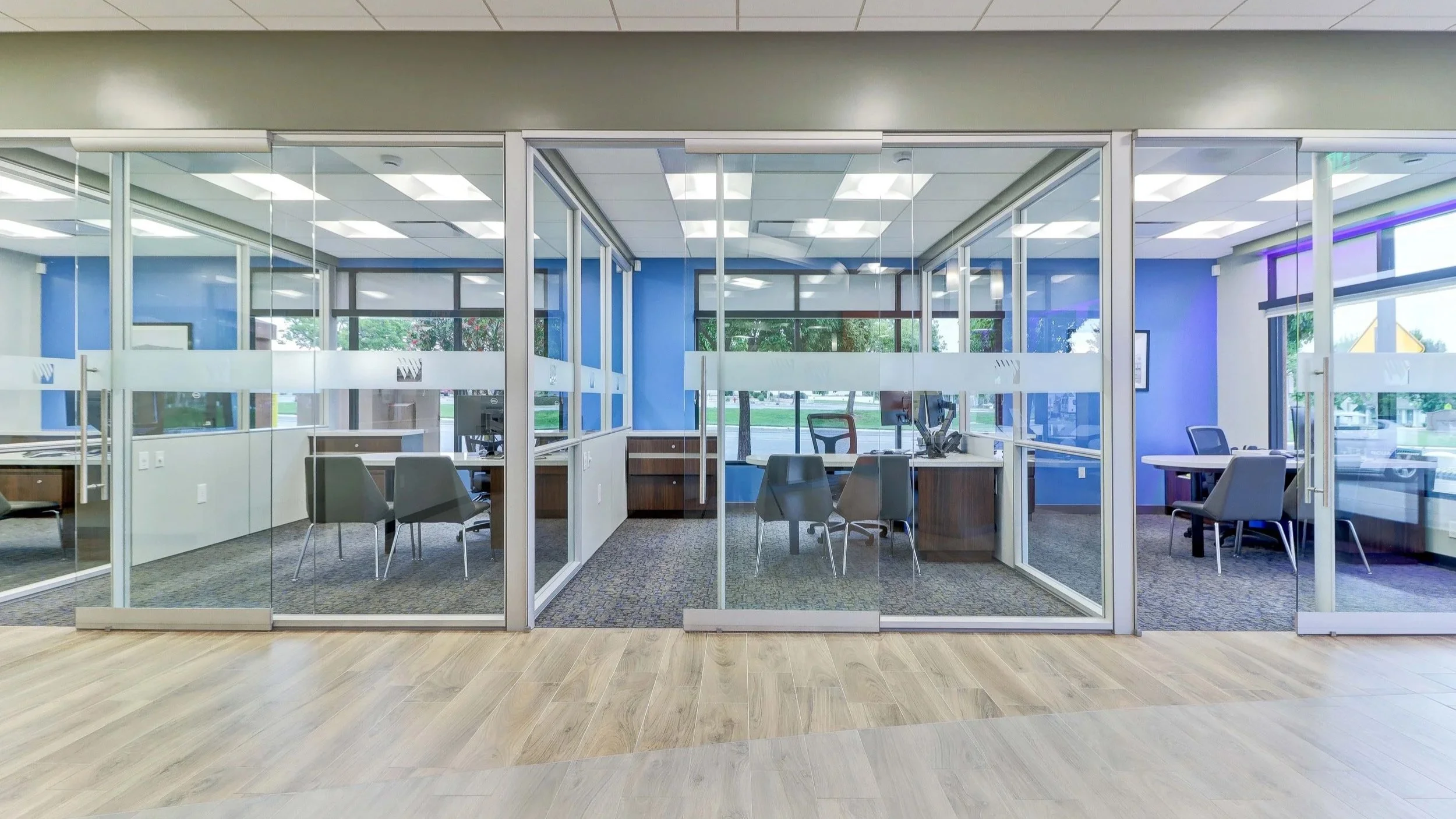 Empty office reception area with glass partition walls, chairs, desks, and large windows displaying trees outside.