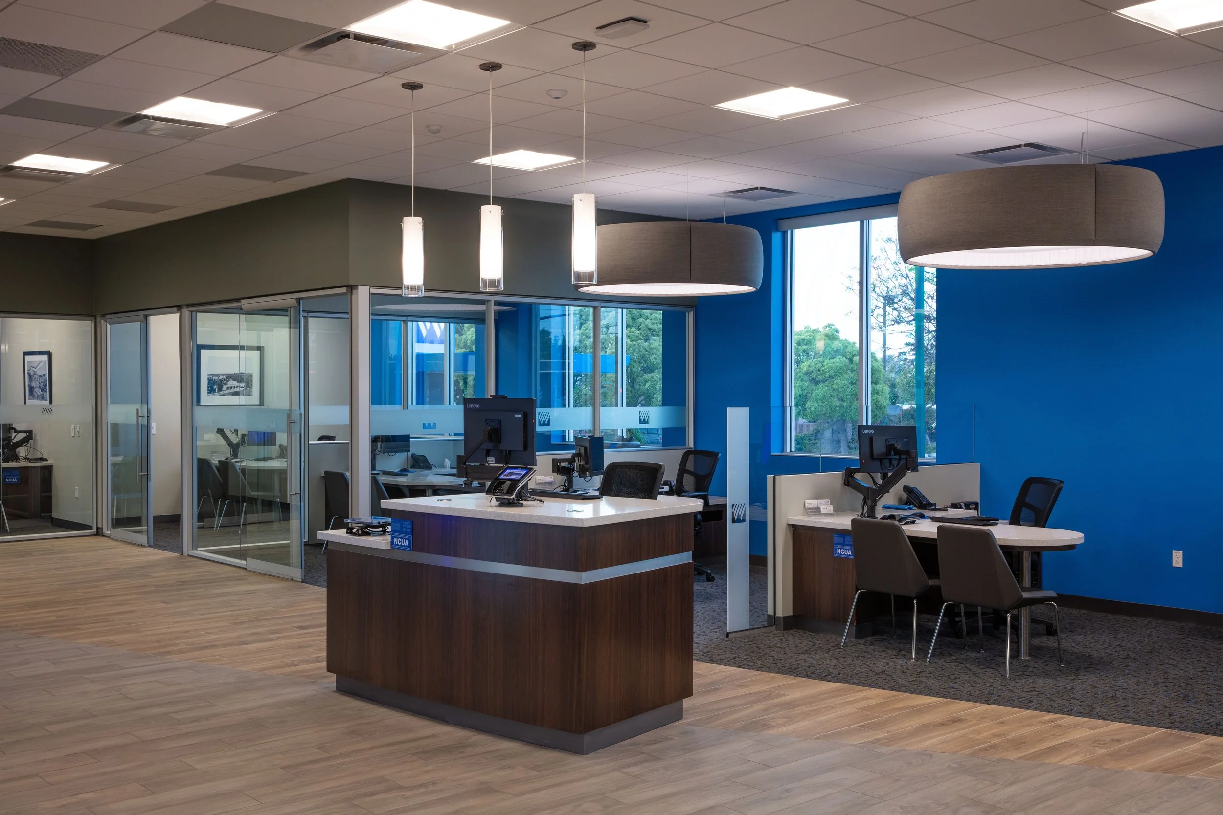 Modern office reception area with a wooden desk, multiple chairs, computer monitors, and office supplies. The background features glass-walled private offices, large windows with a view of trees, and contemporary hanging light fixtures in a professio