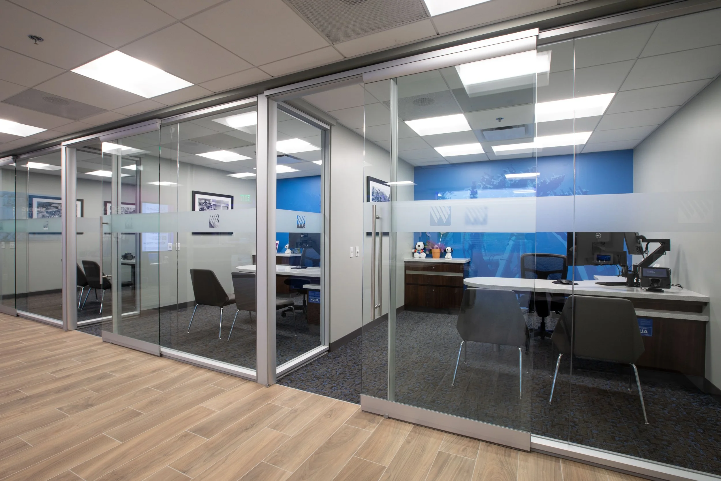 An office conference room with glass walls and sliding doors, interior design includes blue accent walls, framed black-and-white pictures, a desk with two stuffed Snoopy toys, and black chairs, ceiling with recessed lighting, and a wooden floor with 