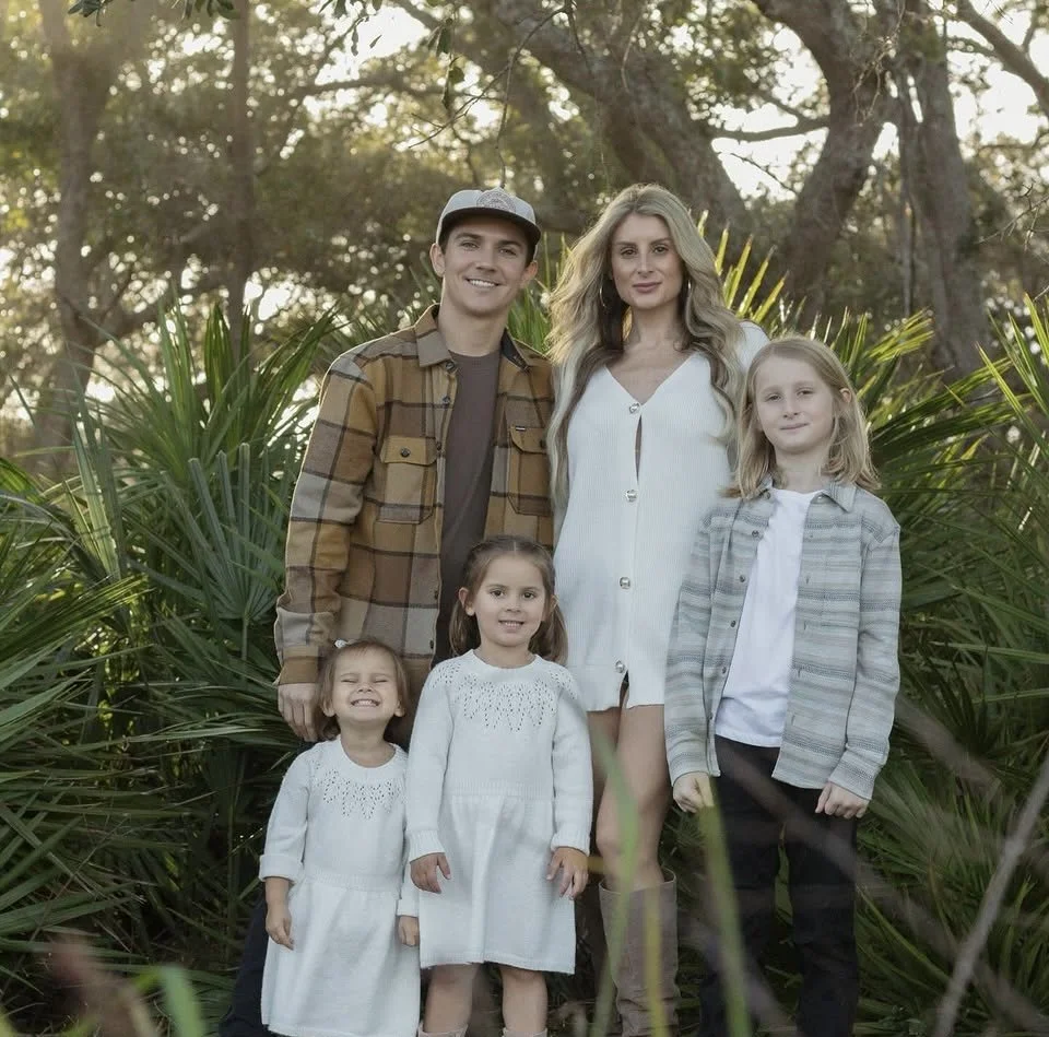 A family of six standing outdoors among trees and shrubbery, smiling at the camera. The group includes two adult males and four young girls, with the adults and children dressed casually.
