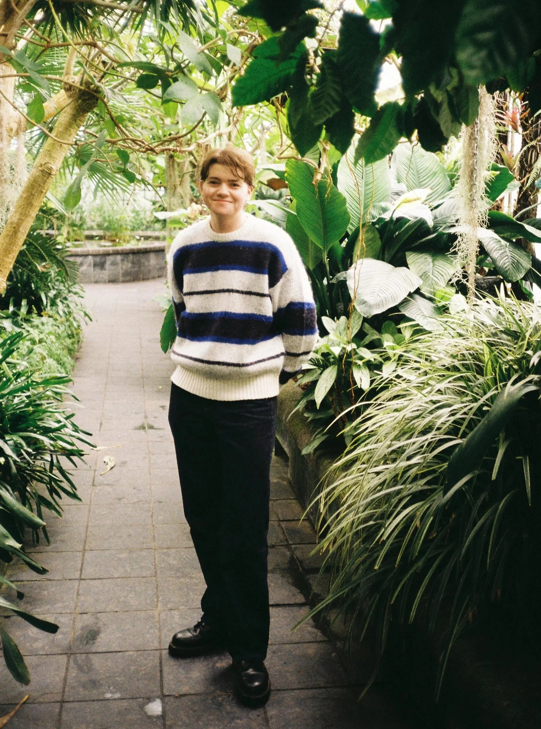 A young person with short, light brown hair stands on a paved path among lush green plants and trees. They are smiling, wearing a cream-colored sweater with dark blue and black horizontal stripes, and black pants and shoes. The background features a brick wall and dense foliage.