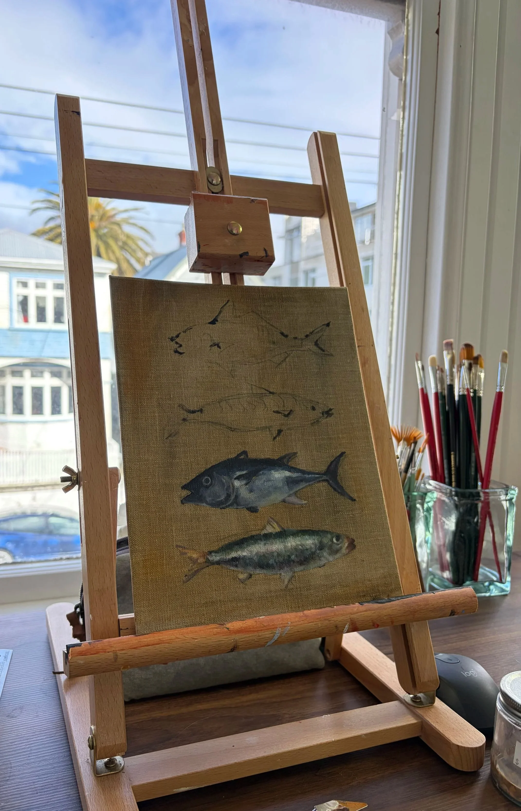 Small painting of three fish on canvas, mounted on a wooden easel, with art supplies nearby.