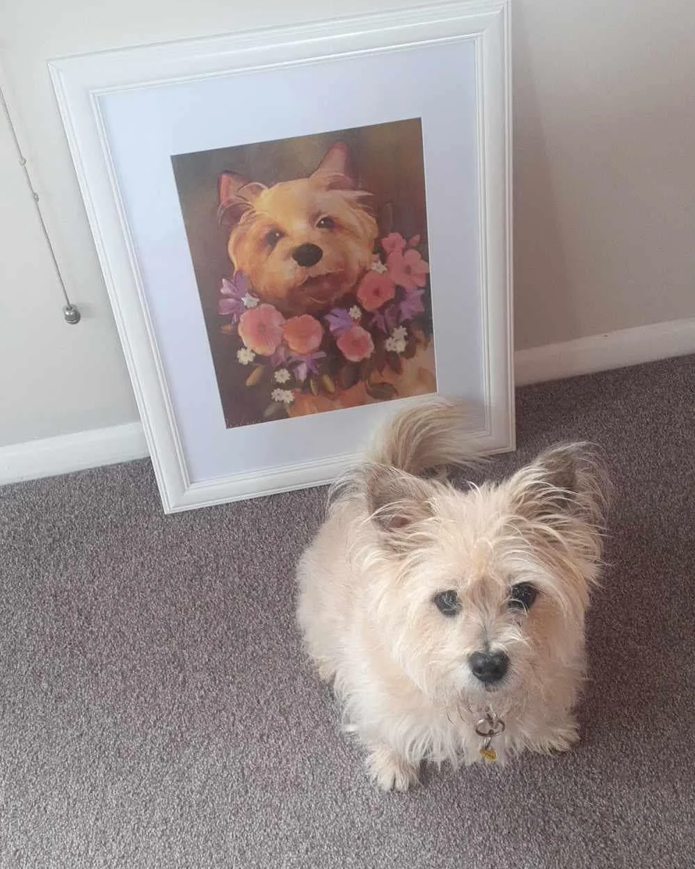 A small white fluffy dog sitting on a gray carpet in front of a framed painting of a dog with a floral wreath around its neck, leaning against a white wall.