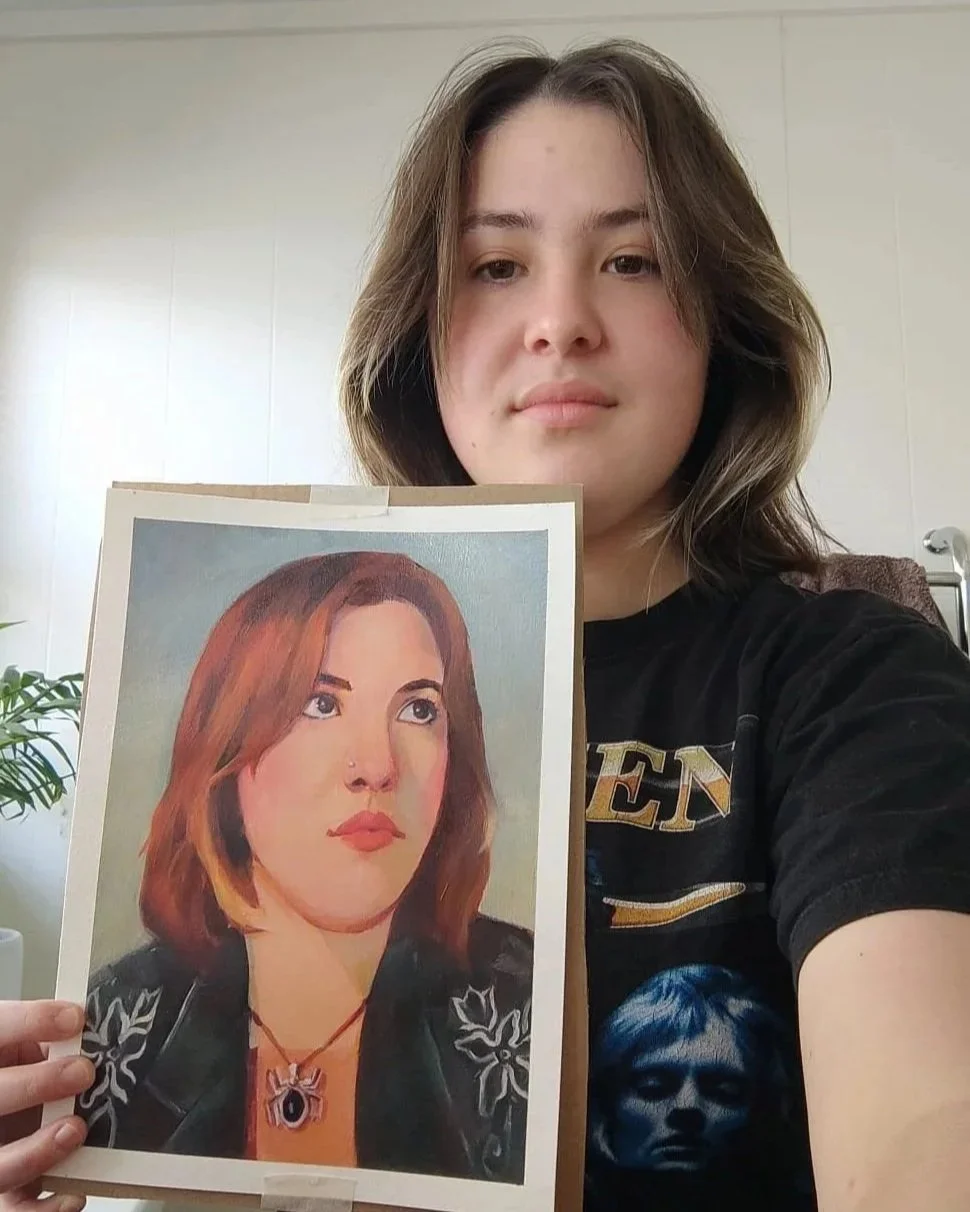 A young woman holding a painted portrait of herself, with medium-length reddish-brown hair and a serious expression.