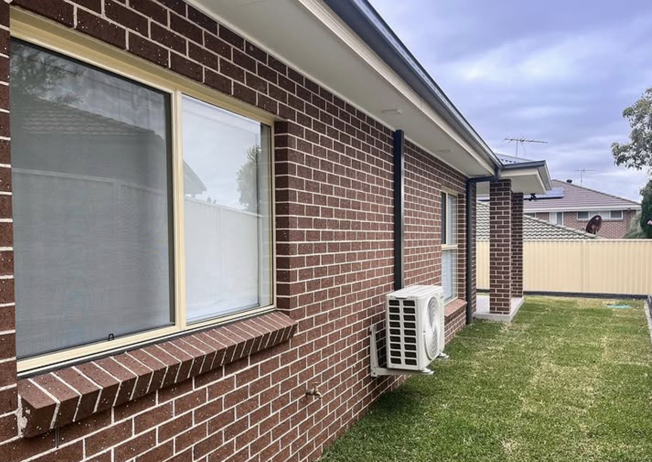 Side view of a brick house with a large window, an air conditioning unit, and a small yard enclosed by a fence.