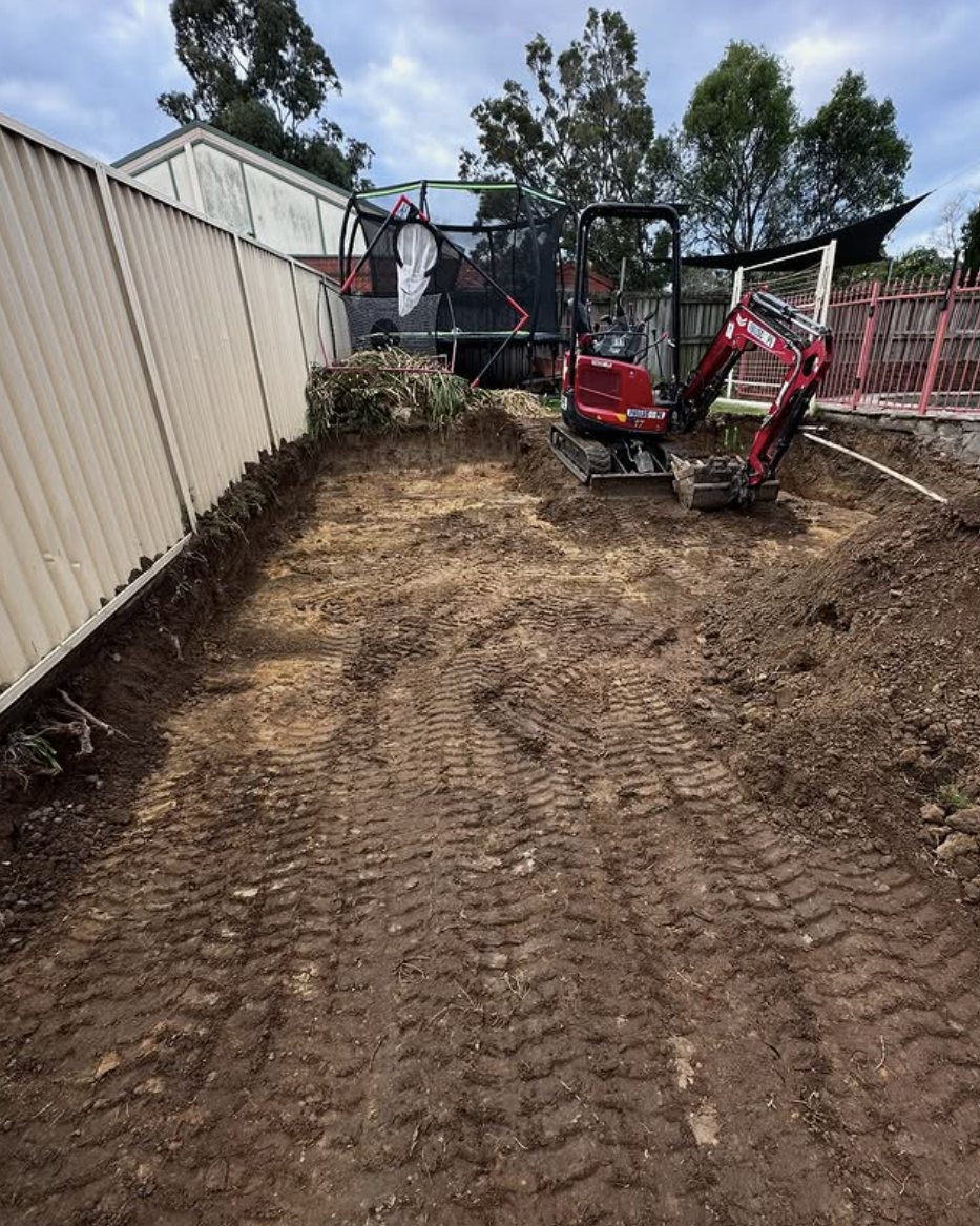 A backyard construction site with a mini excavator and a trampoline in the background. The ground is being prepared for landscaping or foundation work with visible tire tracks.