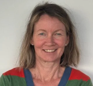 A woman with shoulder-length light brown hair, smiling, wearing a red and green striped sweater, standing against a plain white wall.