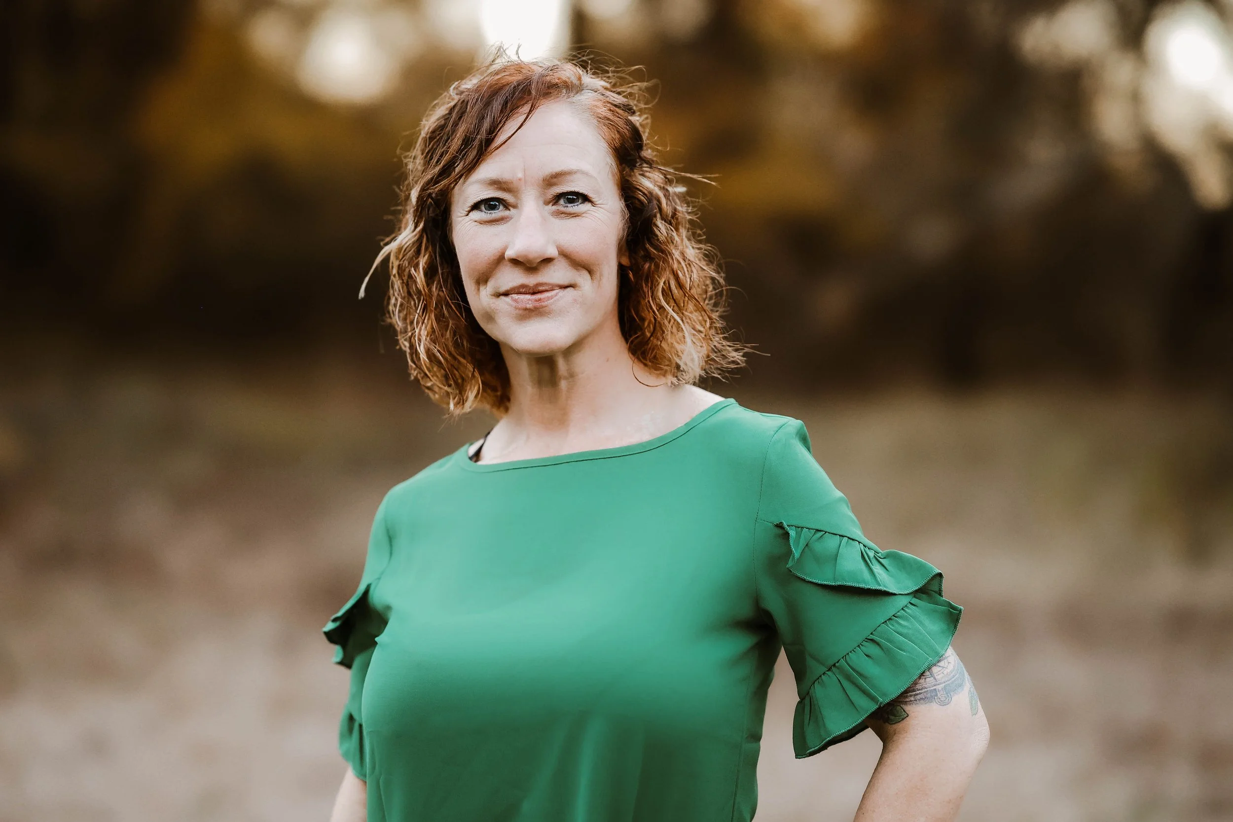 Portrait of a woman with red hair and blue eyes, wearing a green blouse with ruffled sleeves, standing outdoors with blurred trees in the background.