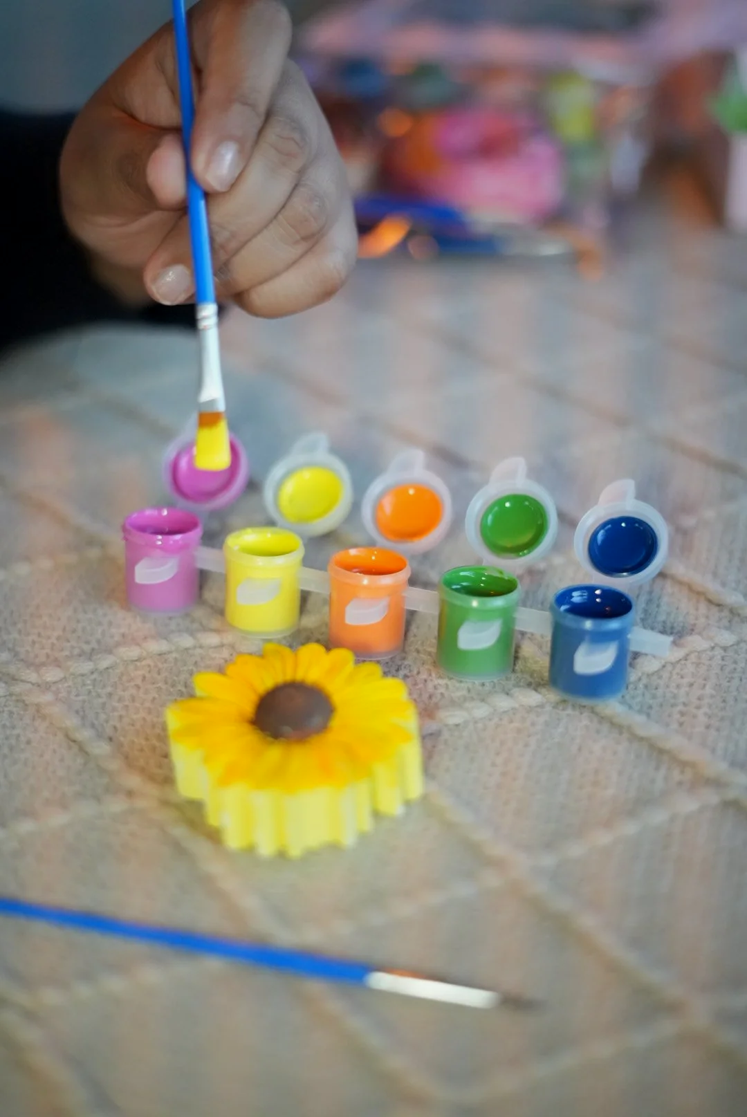 A hand holding a paintbrush with yellow paint, dipping into small open containers of colorful paints arranged on a fabric surface, with a sunflower in the foreground and other art supplies in the background.