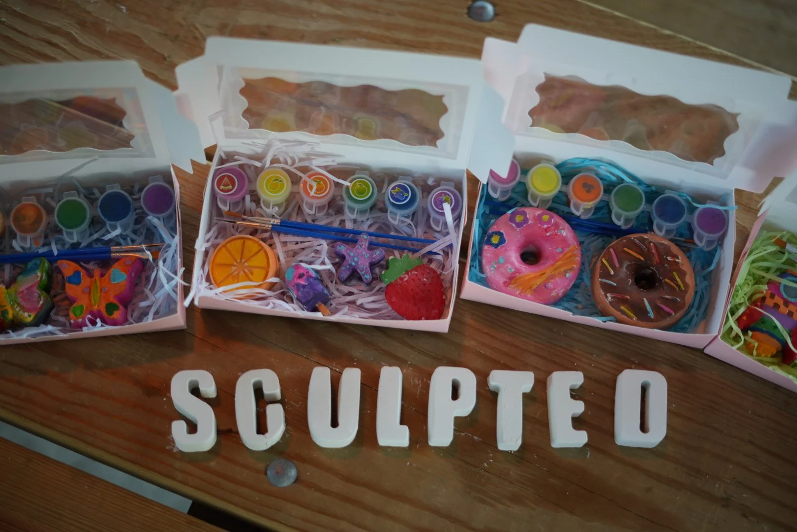 Assorted painted rock crafts in decorative boxes, including butterfly, orange slice, starfish, strawberry, pink donut, and chocolate donut, displayed on a wooden surface with the word "SCULPTED" spelled out in white letters.