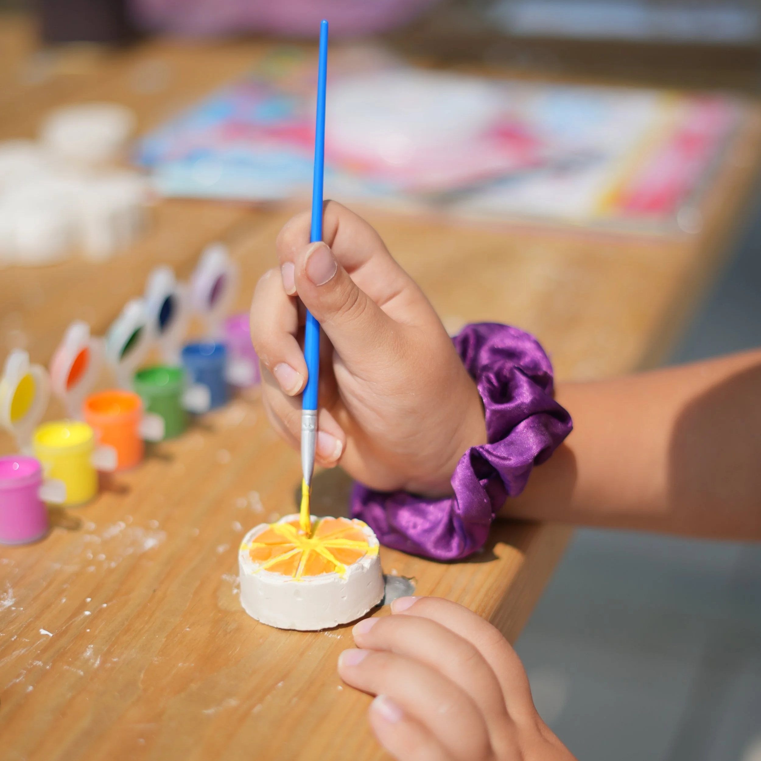 A child holding a blue craft stick, in front of a craft project with orange and lemon slice design, on a wooden table, with colorful paint bottles in the background.