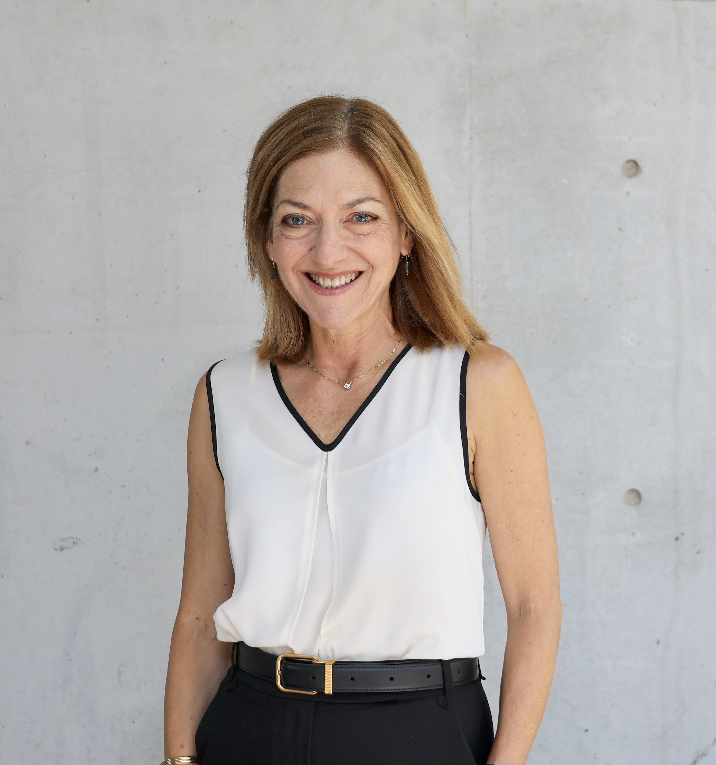 Close-up of a smiling woman with light skin, light blue eyes, and shoulder-length reddish-brown hair, wearing a sleeveless white top with black trim, a black belt, gold jewelry, and standing against a concrete wall background.