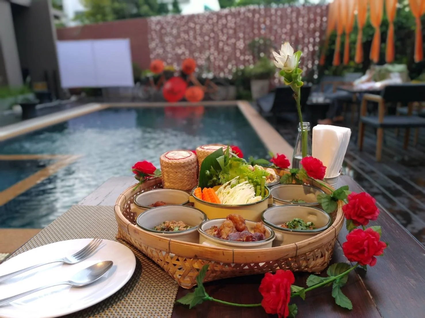 An outdoor dining setup near a pool with a basket of food containing various dishes, a plate with a fork and spoon, and a vase with a white flower, decorated with red flowers, on a wooden table.