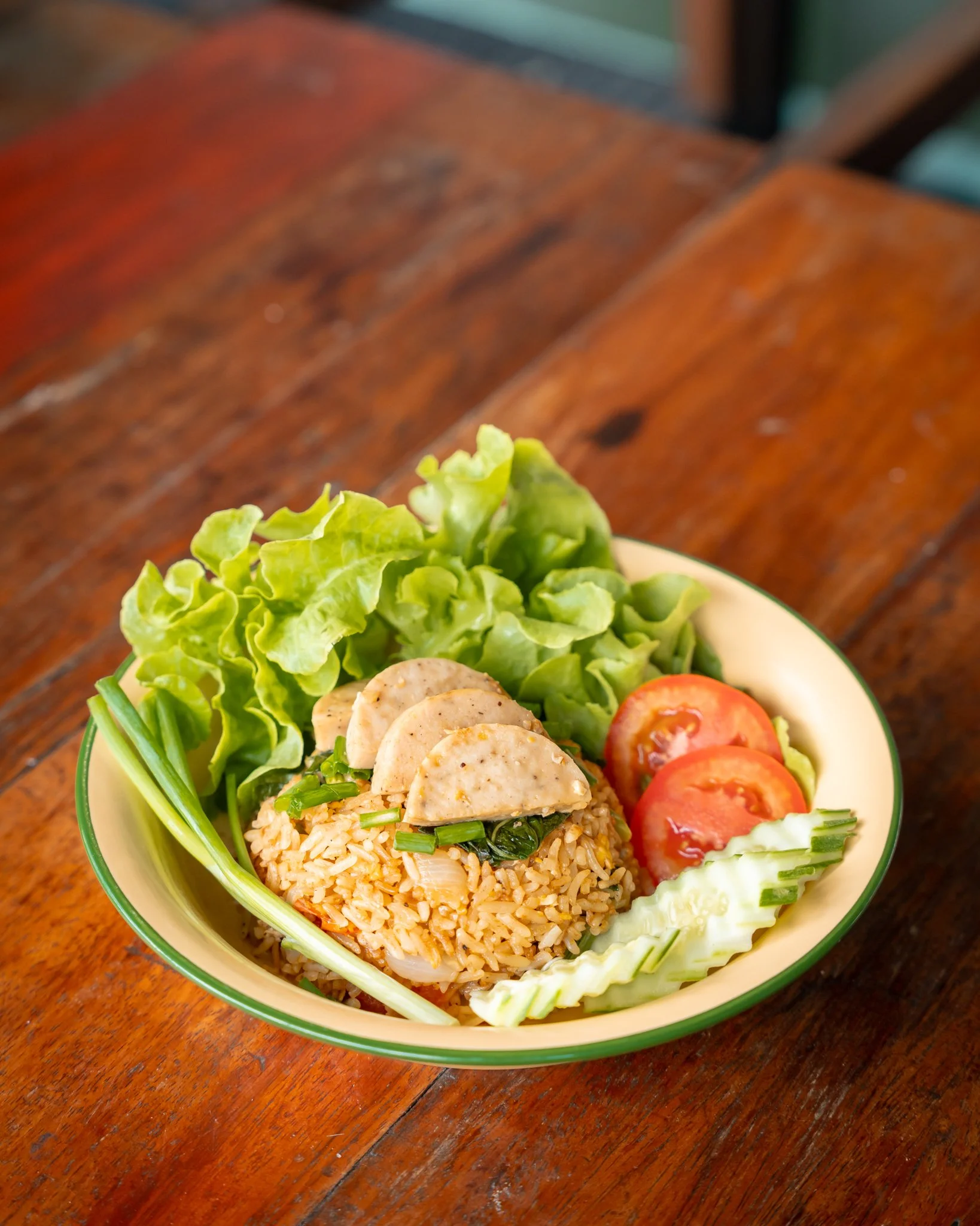A bowl of fried rice with sliced chicken, fresh lettuce, tomato slices, cucumber slices, and green onions on top. The bowl is sitting on a wooden table.