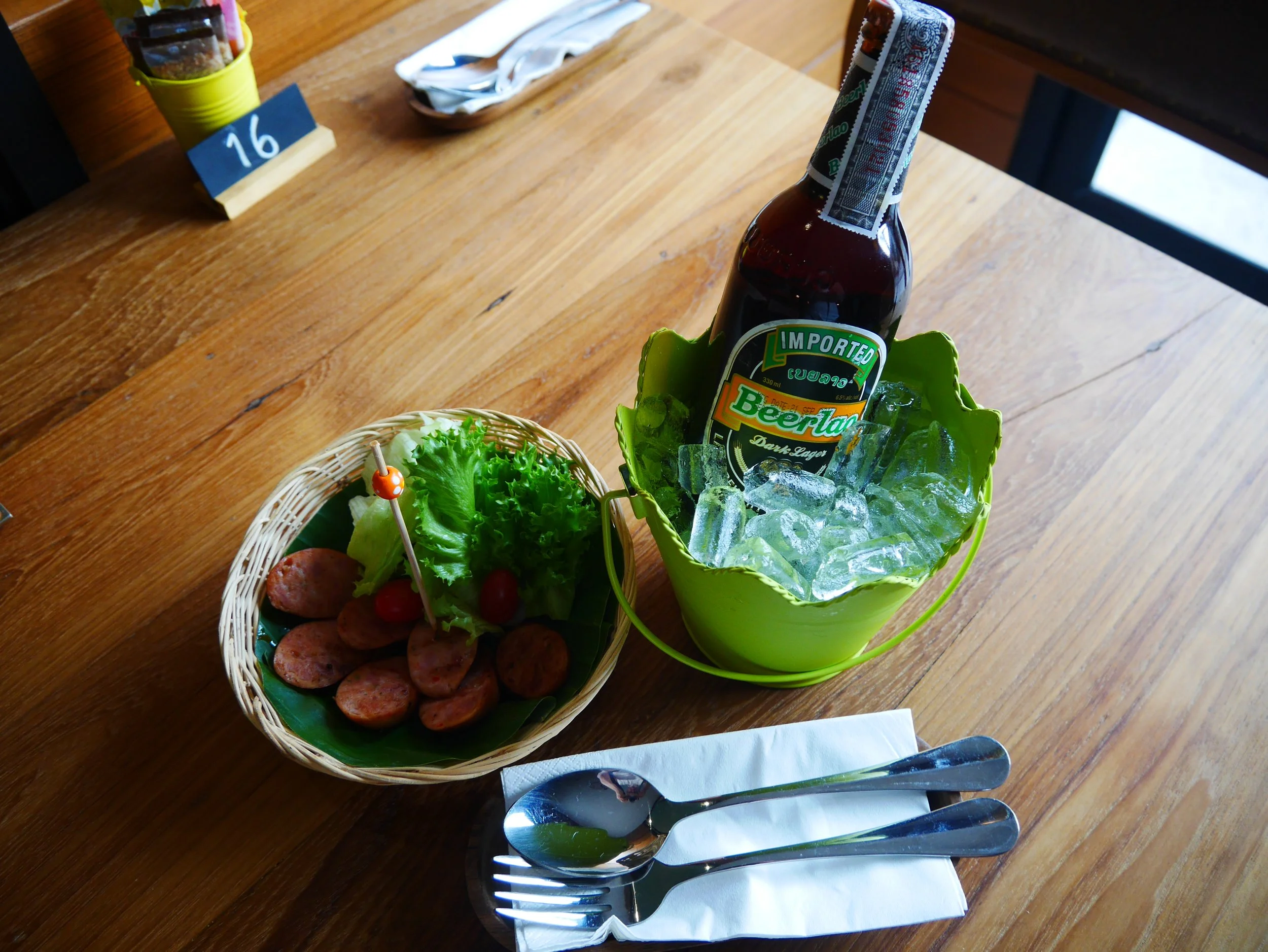 A wooden table set with a basket of sliced sausages, a lettuce garnish, and a drink in a green bucket filled with ice, surrounded by utensils and a napkin.