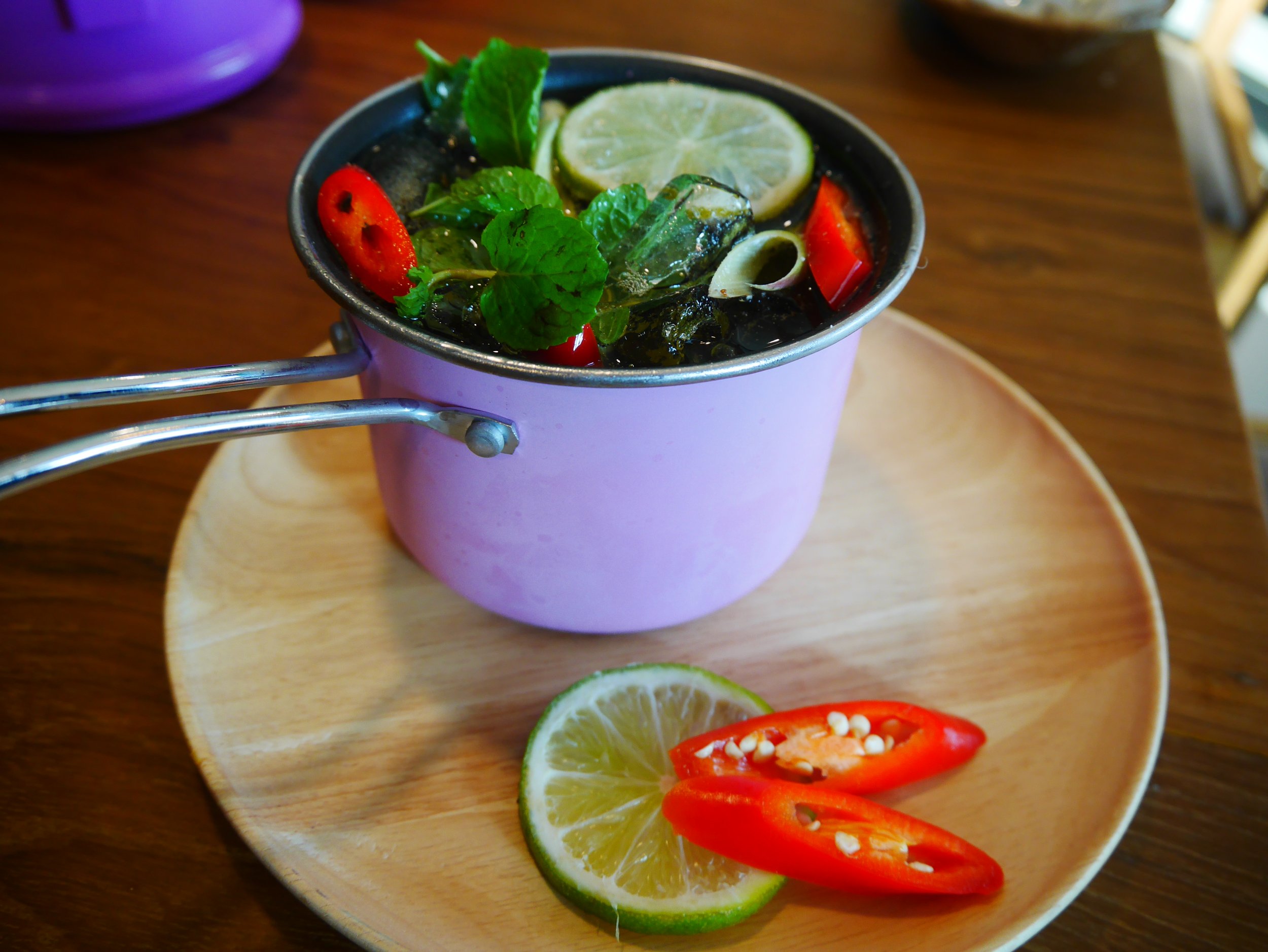 Lemon slices, red chili slices, and fresh mint leaves on a small pink cup, sitting on a wooden plate.