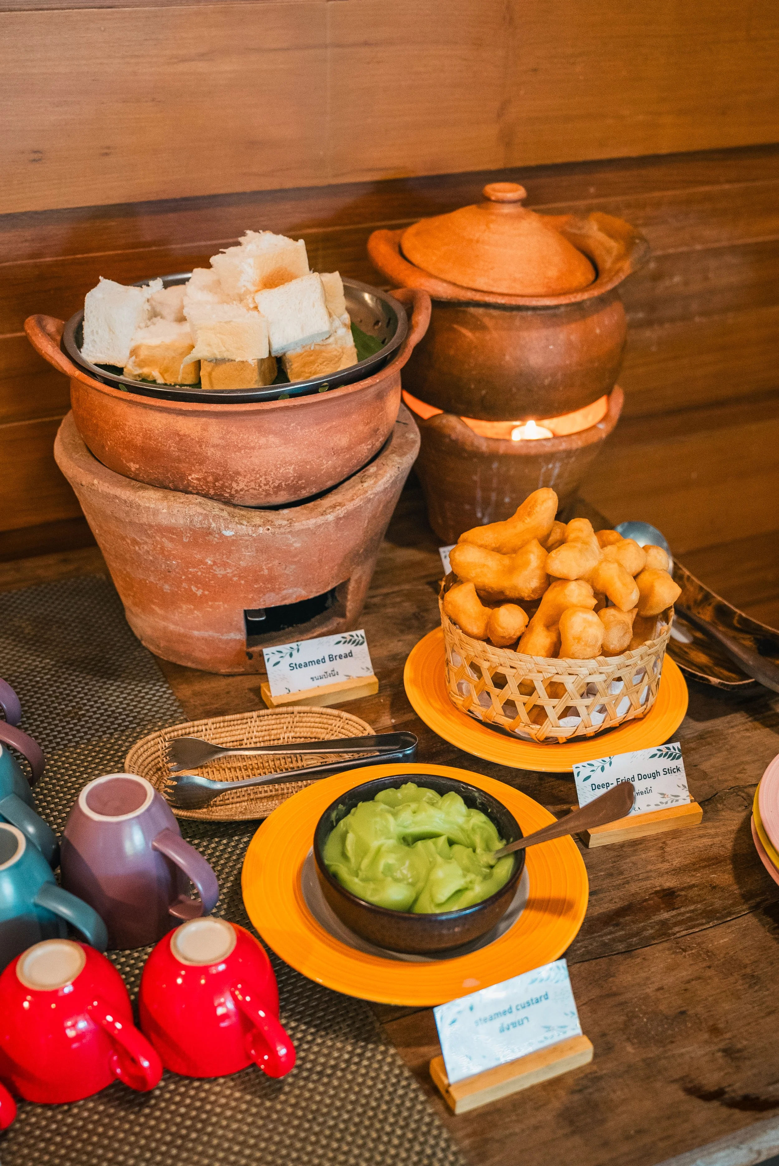 A spread of breakfast foods on a wooden table, including steam bread, deep-fried dough sticks, steamed custard, and green custard, with colorful cups and small plates.