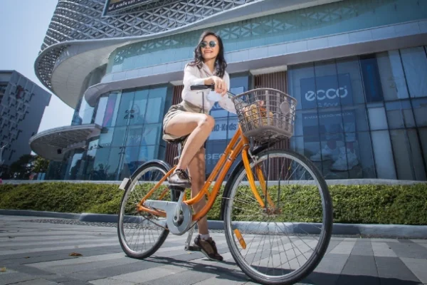 A woman wearing sunglasses and white blouse riding an orange bicycle with a basket in front, in front of a modern shopping mall.