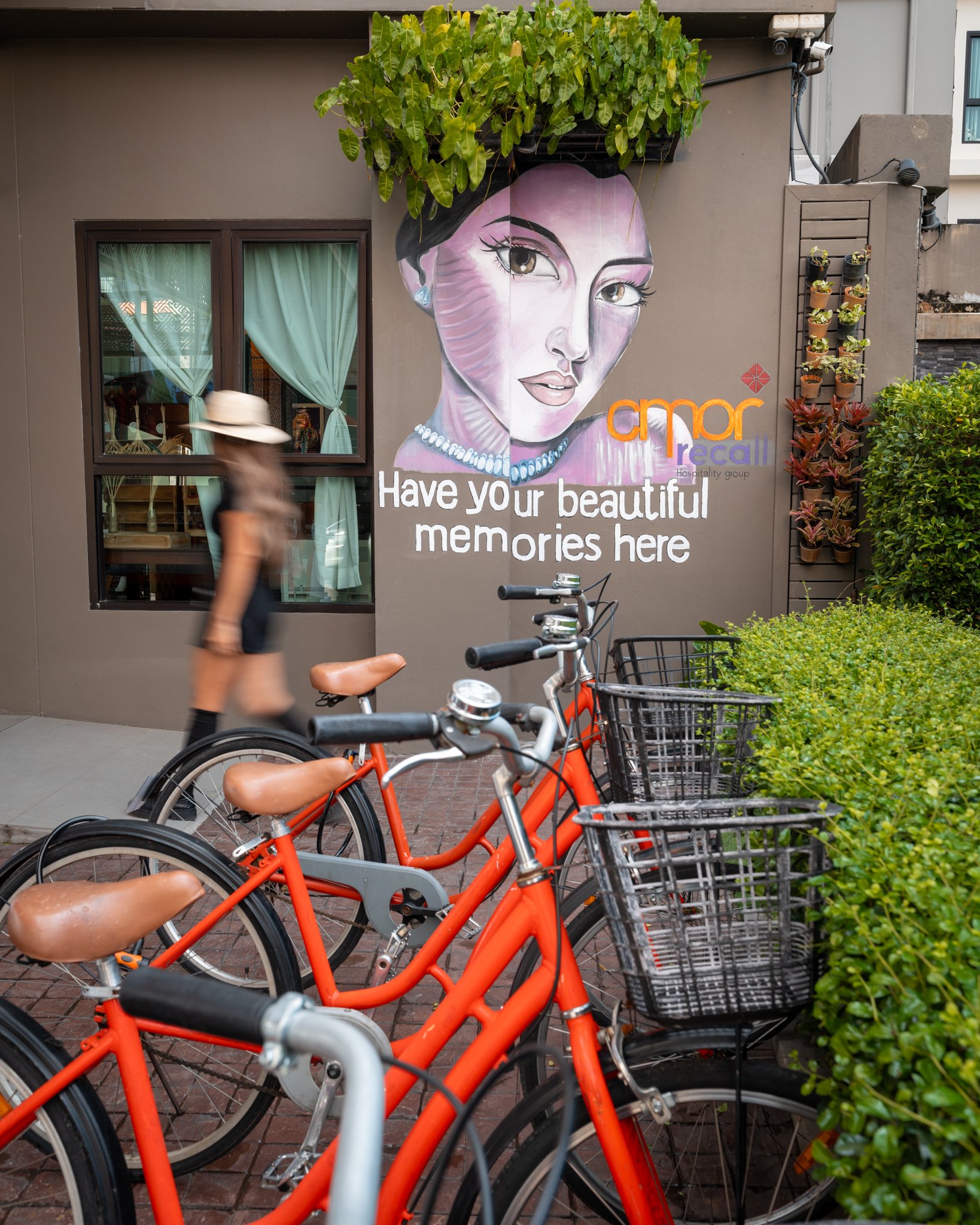 Bright orange bikes parked in front of a building with a large colorful mural of a woman's face, with text that reads 'Have your beautiful memories here' and a bicycle with a basket at the front.