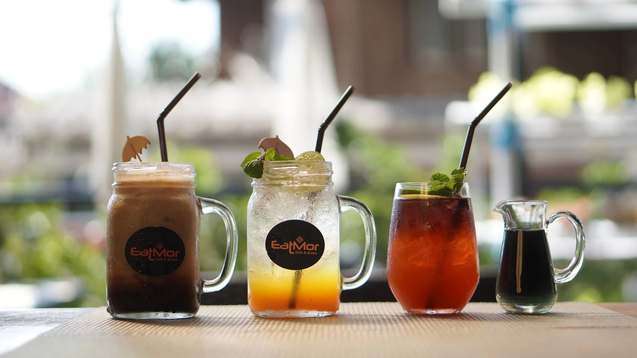 Three colorful beverages in glass jars with handles, garnished with mint and other fruits, on a table in a cafe setting.