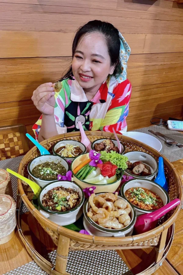 Woman smiling and holding a piece of food at a restaurant, with a large spread of various dishes in bowls and a basket in front of her.