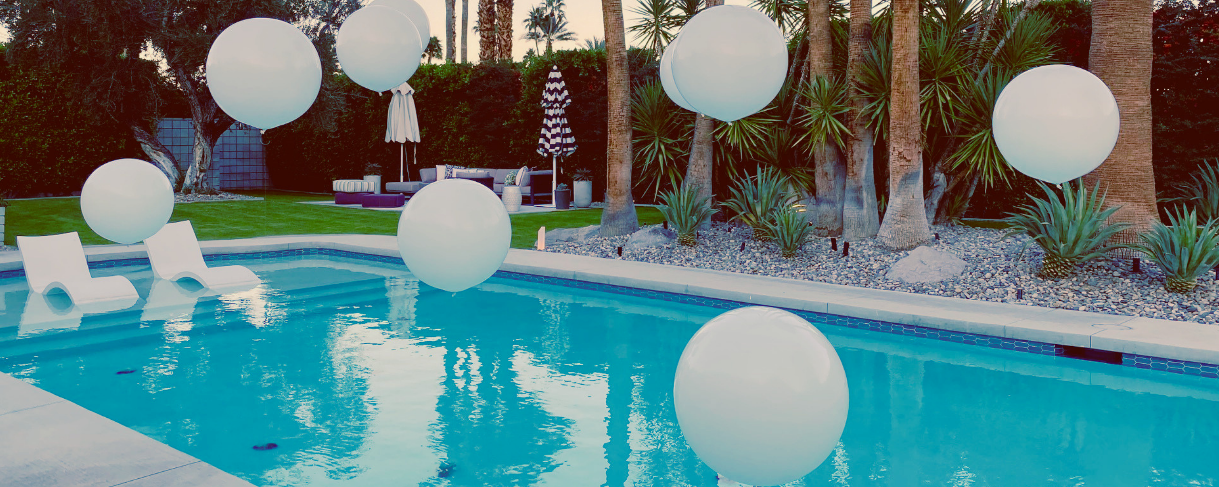 A backyard swimming pool with floating white spheres, two white pool chairs, surrounded by palm trees and outdoor furniture with umbrellas.