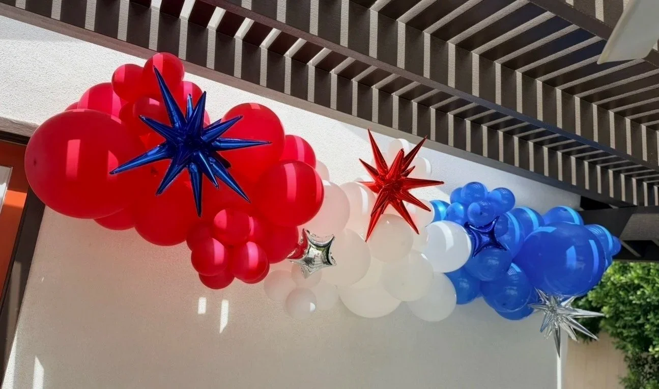 Colorful balloon garland with red, white, and blue balloons and star-shaped balloons in red, blue, and silver hanging outside during daytime.
