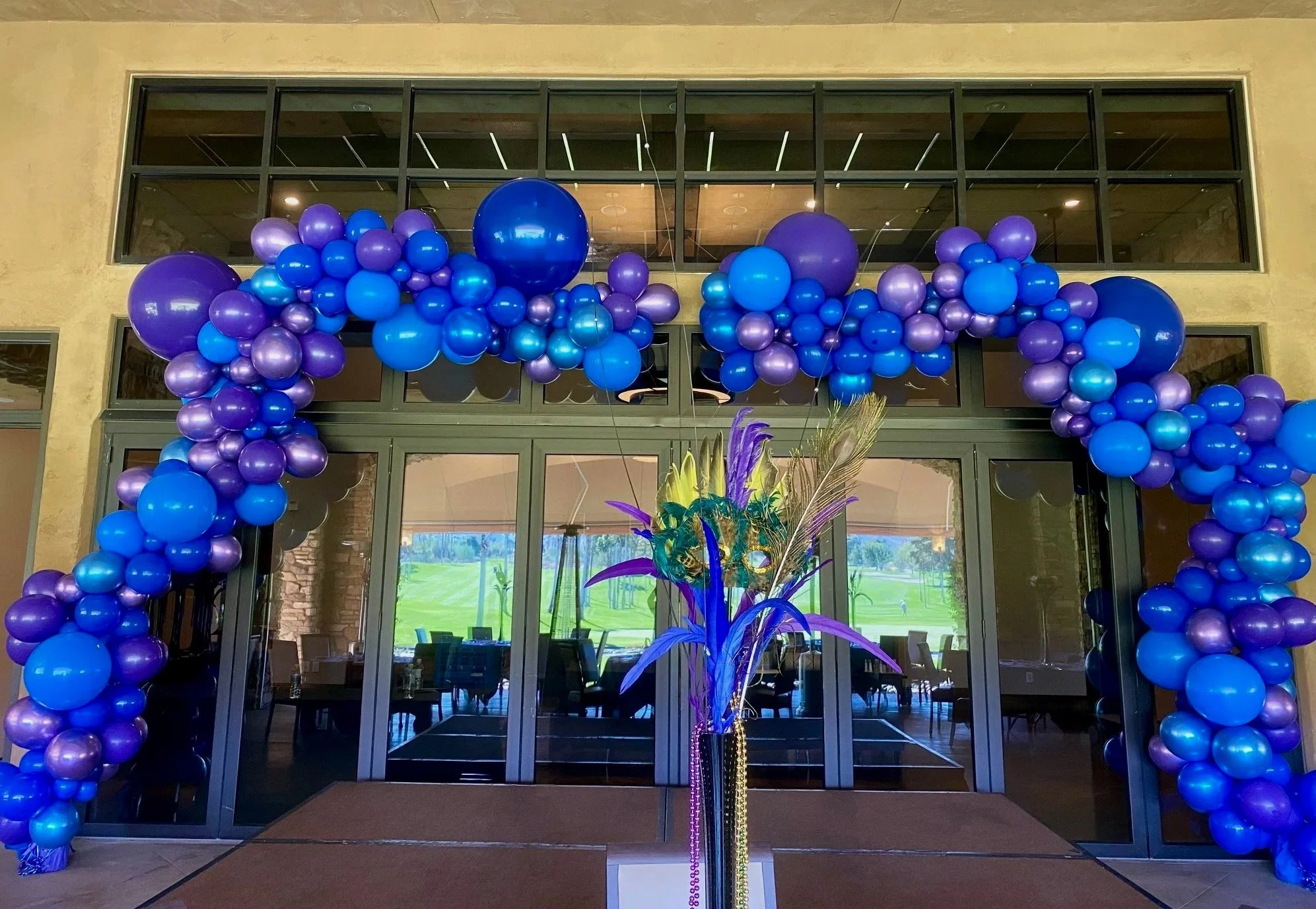 An entrance decorated with blue and purple balloons arranged on both sides and above the doorway, with a large floral arrangement featuring purple and yellow feathers in the foreground, inside a building with large glass windows showing a green outdo