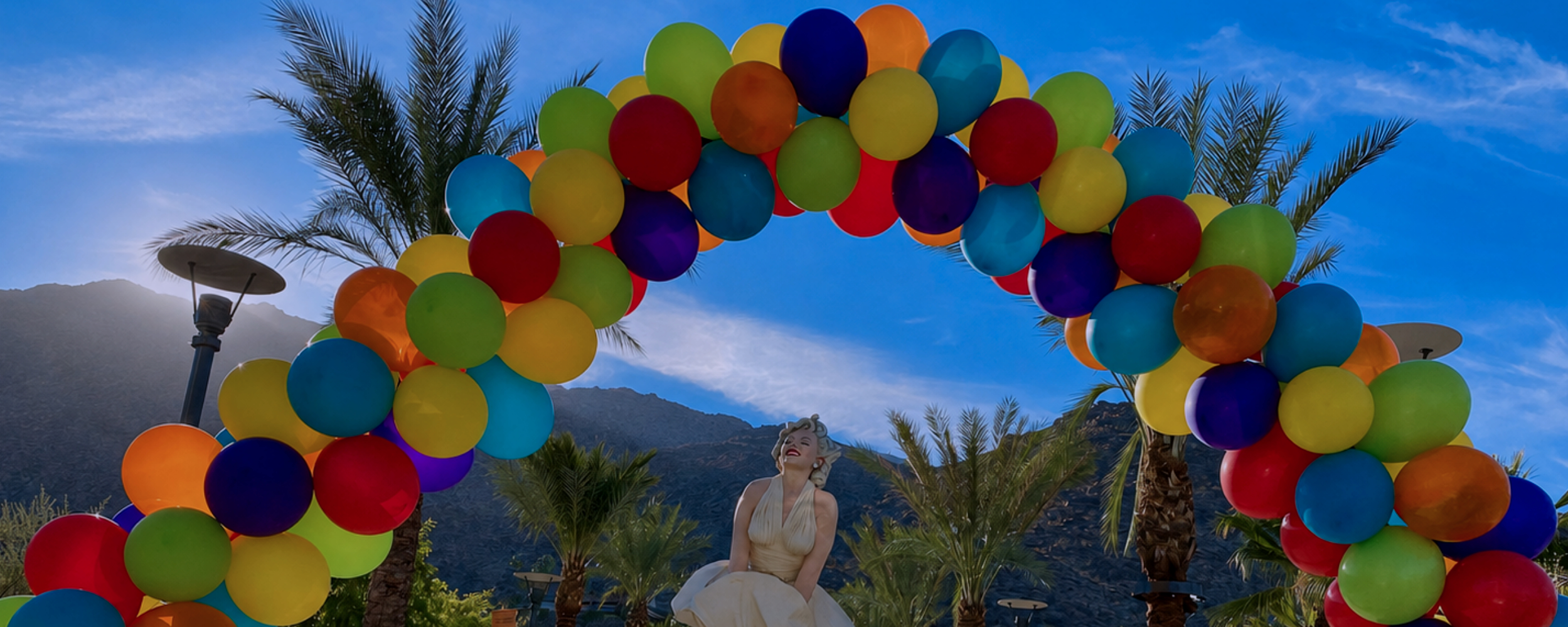 Colorful balloons arranged in an arch, with palm trees and mountains in the background under a blue sky, and a Marilyn Monroe woman statue beneath in Palm Springs, CA.