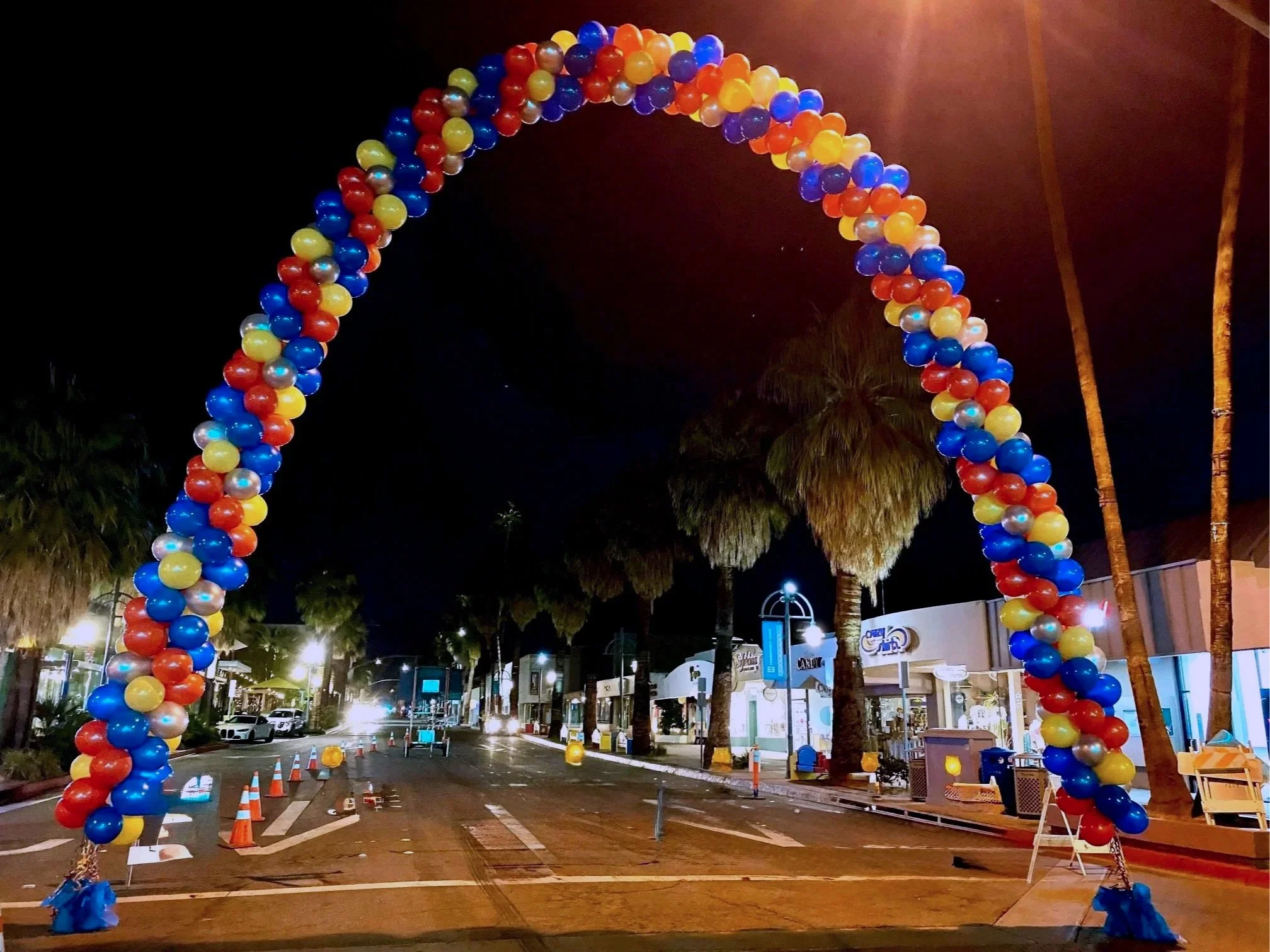 A street decorated with a large arch made of colorful balloons at night. There are palm trees, shops, and cars in the background, with street lights illuminating the scene.
