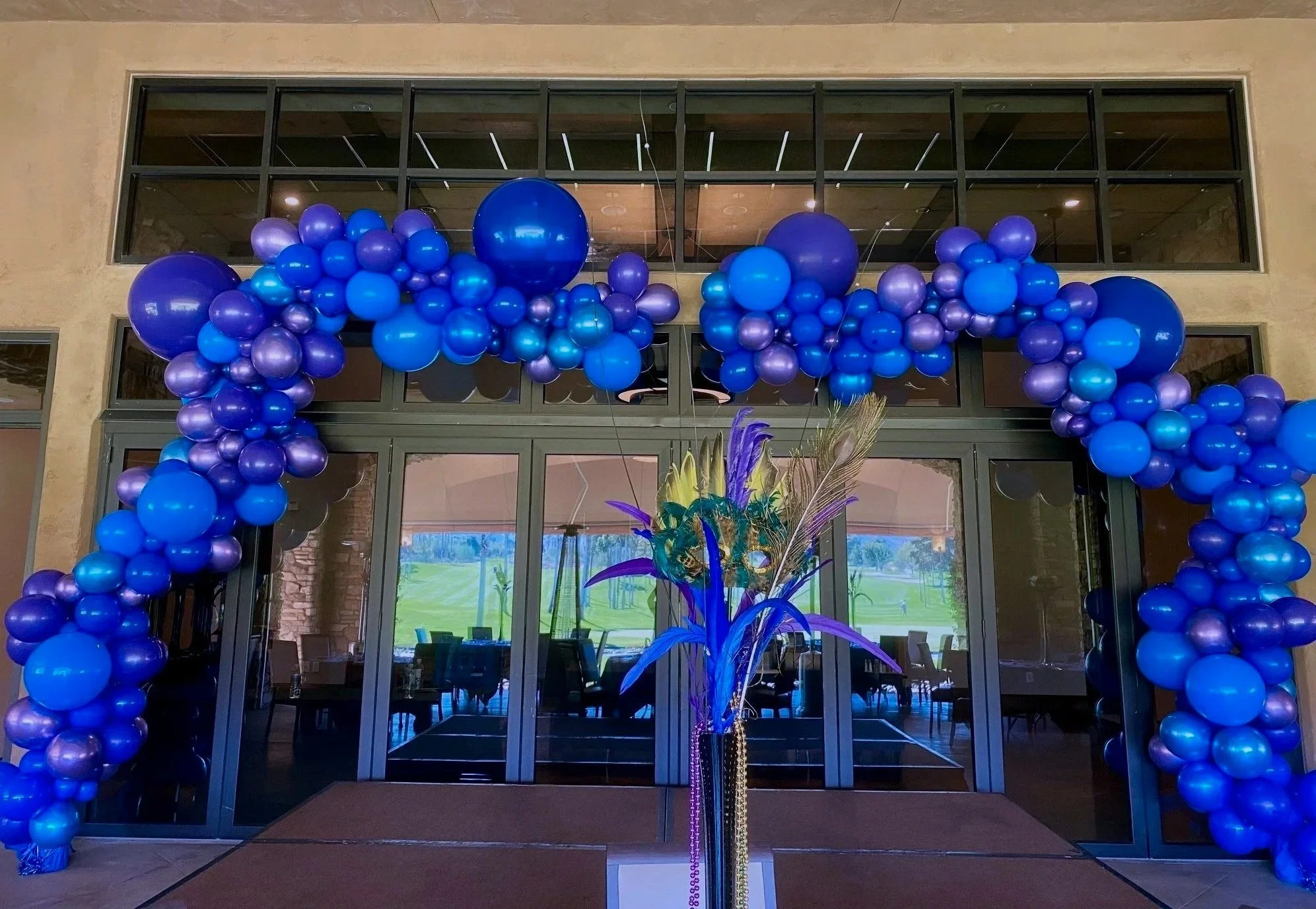 Decorative balloon arch in shades of blue and purple framing a glass entrance door with a tropical flower arrangement in front.