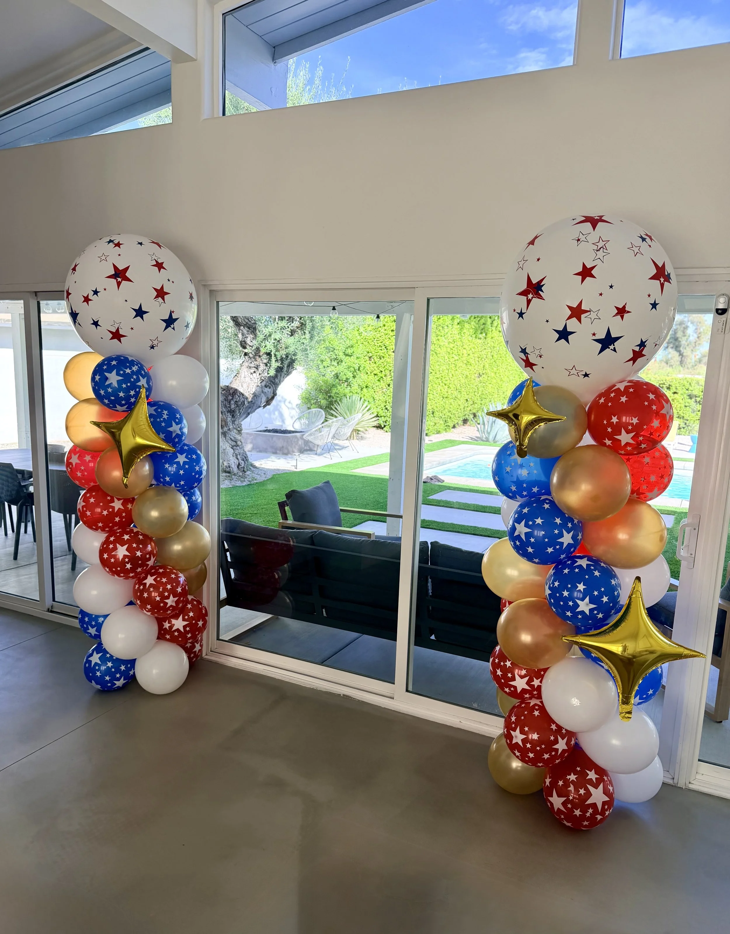 Decorative balloon columns with red, white, blue, and gold balloons, featuring star patterns and gold star-shaped balloons, placed inside a house near a large glass sliding door with outdoor greenery visible.